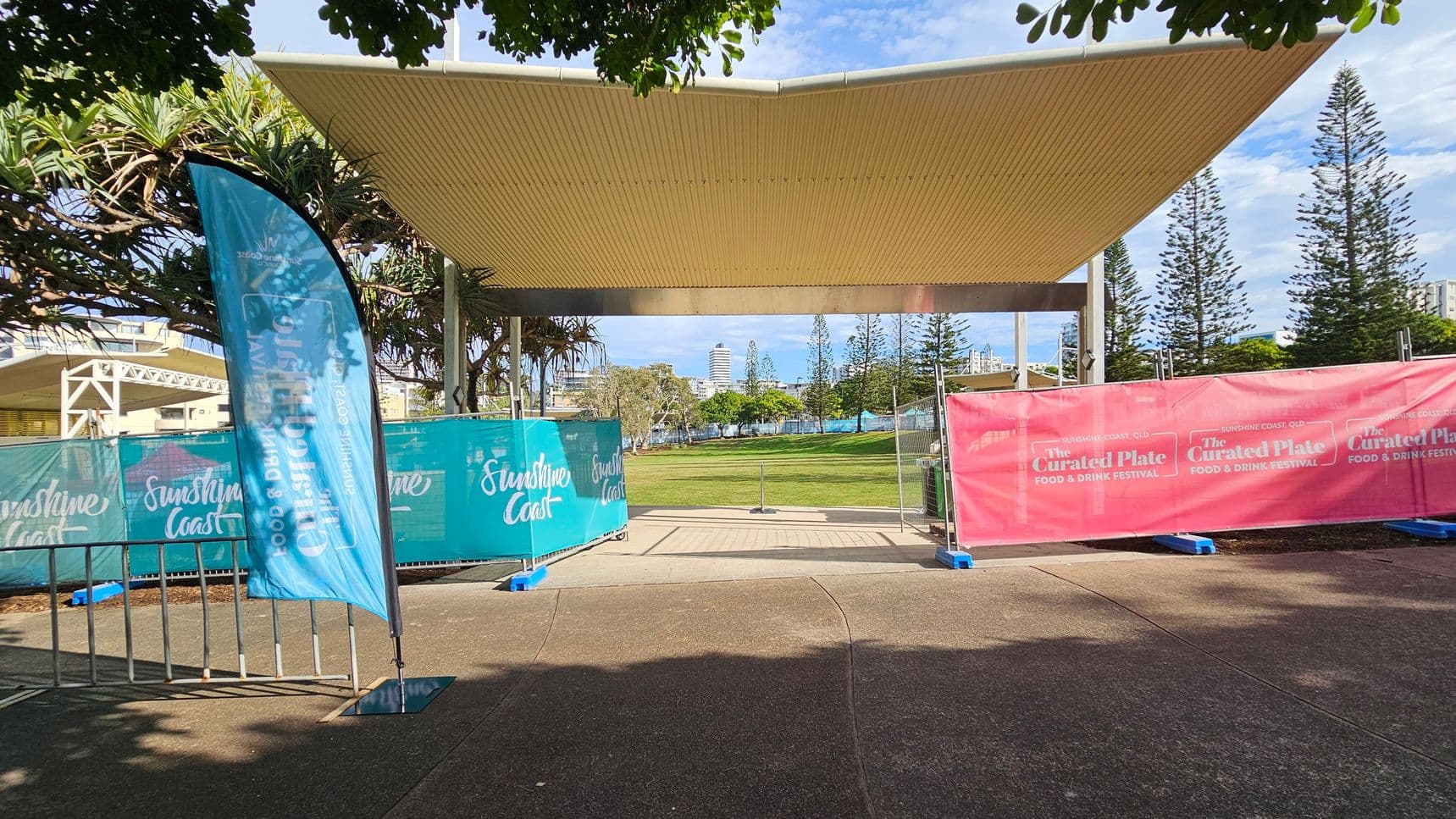 A covered outdoor area with a corrugated roof. In the foreground, there are blue and pink banners with "Sunshine Coast" and "The Curated Plate Food & Drink Festival" written on them. A blue flag stands on the left. The ground is paved, and there are trees and a grassy area in the background. Tall buildings and more trees are visible in the distance.