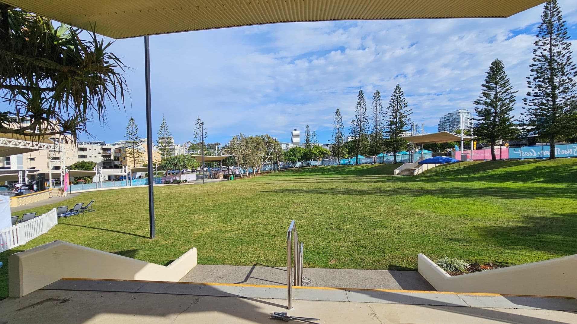 A grassy park area with a few scattered trees and a row of tall, slender trees in the background. On the left, there are buildings and a few lounge chairs near a white fence. In the centre, a metal handrail leads down steps to the grass. On the right, more trees and a covered structure are visible. The foreground has a concrete surface, and the ceiling above is partially visible with some hanging plants.