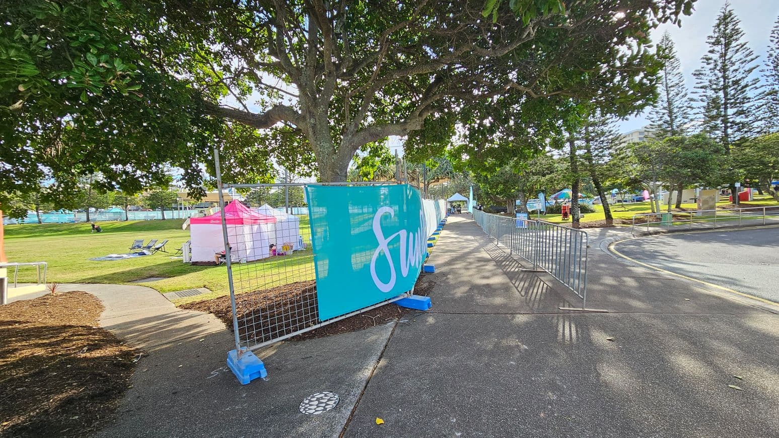 A pathway lined with metal barriers on the right, with a large tree providing shade. A blue banner with white text is attached to the fence. On the left, a grassy area with pink and white tents, deck chairs, and a few people sitting. In the background, more trees and a building are visible. The ground is paved, and the area is well-lit with sunlight filtering through the leaves.