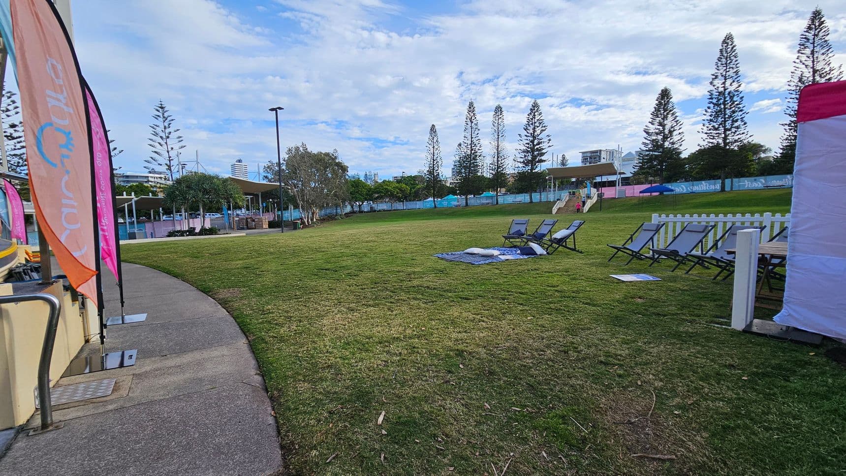 A grassy area with several deck chairs and a blue blanket on the right. A white picket fence is nearby. On the left, there are colourful flags along a concrete path. In the background, tall trees and a few buildings are visible, along with a covered structure and a person walking up steps.