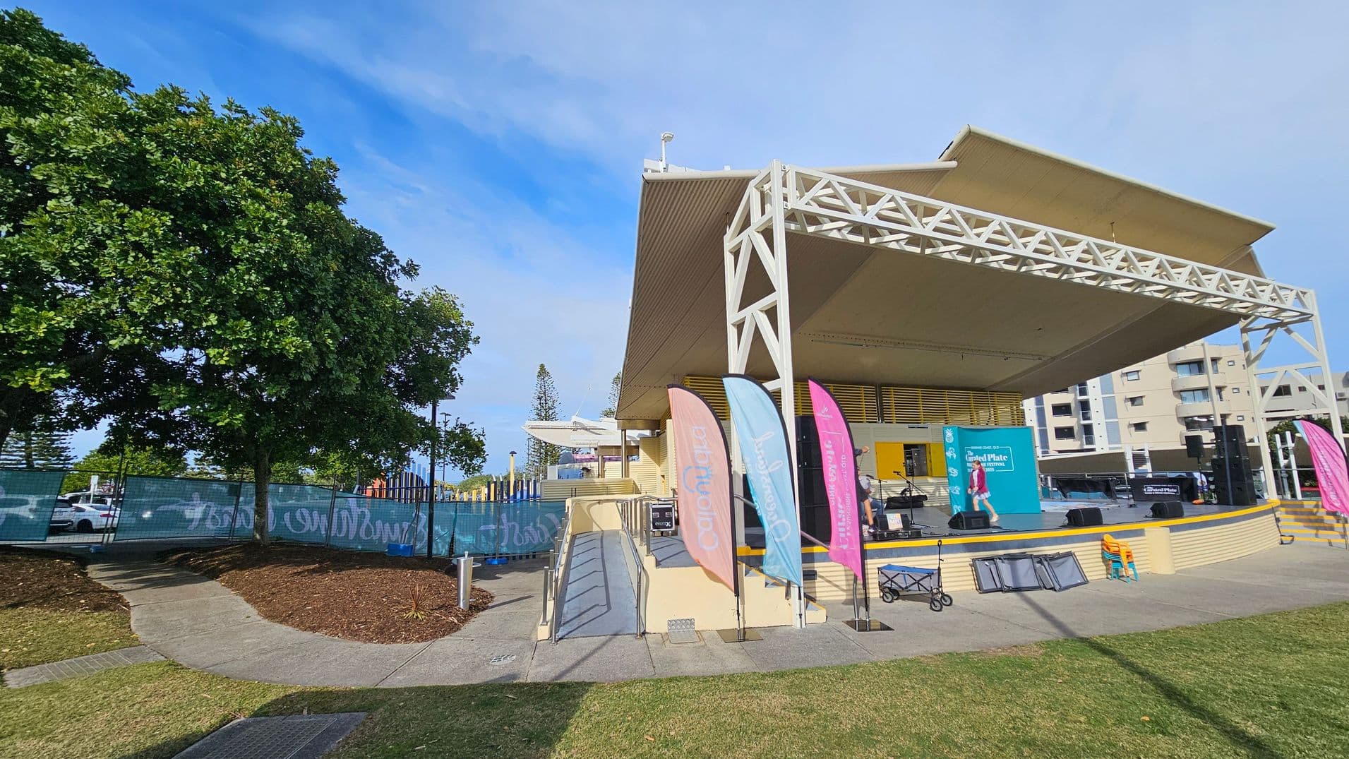 A stage with a large, white, triangular roof structure on the right. Three colourful flags are positioned in front of the stage. A ramp leads up to the stage, with a few musical instruments visible. On the left, a large tree provides shade over a paved path. Behind the tree, a fence with graffiti separates the area from parked cars. In the background, there are several multi-storey buildings. The ground is a mix of grass and paved surfaces.