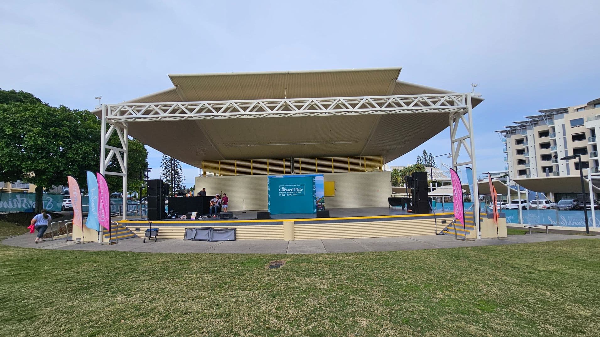 An outdoor stage with a large, white, triangular roof structure. In the centre, a blue sign with text is displayed. On the stage, a few people are setting up equipment. To the left, colourful vertical banners are positioned near a small set of stairs. In the background, there are trees and a multi-storey building. The foreground features a grassy area leading up to the stage.