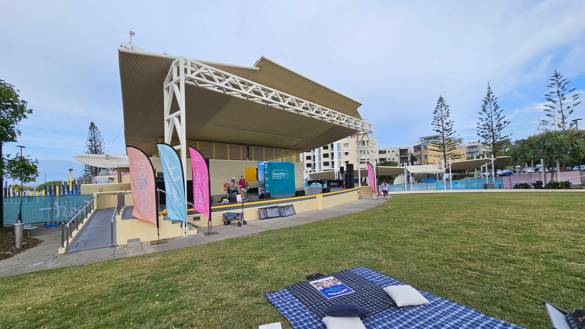 A large outdoor stage with a beige roof and lattice framework is on the left. Colourful flags are positioned in front of the stage. Musicians are performing on stage, with a blue sign behind them. In the foreground, a blue and white checkered picnic blanket with a magazine and cushions is spread on the grass. To the right, there are tall trees and several buildings in the background. A pathway with railings leads up to the stage.
