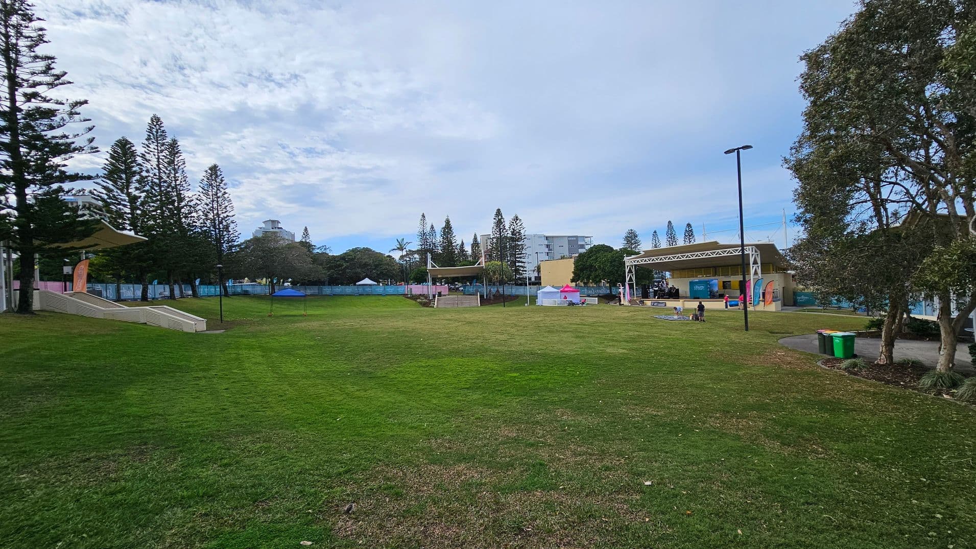 A grassy park area with a stage on the right, featuring a few people and colourful banners. On the left, there are tall trees and a small structure with a canopy. In the background, buildings and more trees are visible. A path with a couple of bins is on the right, and the sky is overcast.
