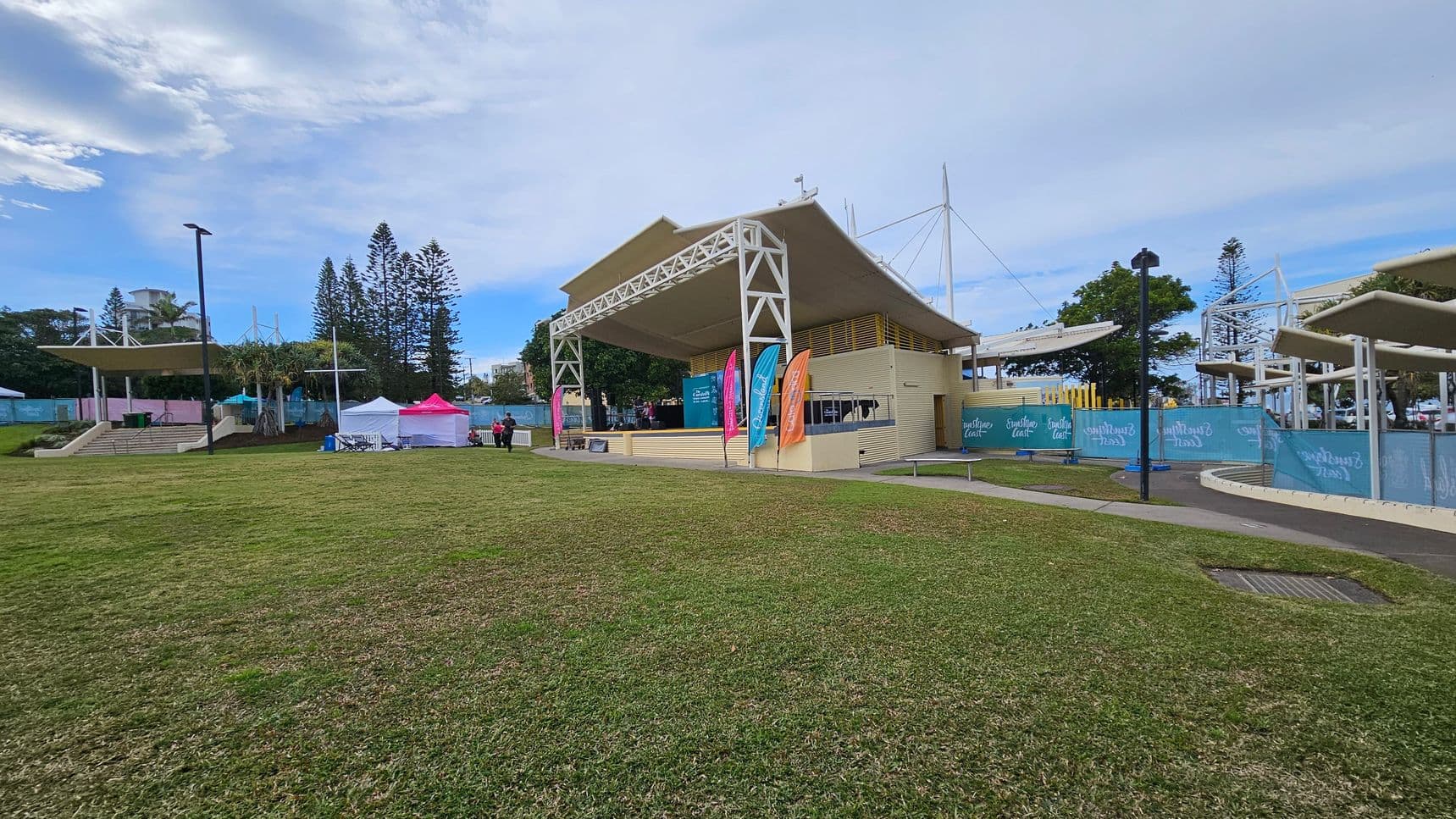 A grassy area in the foreground leads to a stage structure in the centre with a beige roof and white supports. Colourful flags are displayed in front of the stage. To the left, there are two small tents, one white and one pink, with a few people nearby. In the background, tall trees and a few buildings are visible. On the right, there are more structures with white roofs and blue fencing. The sky is overcast.