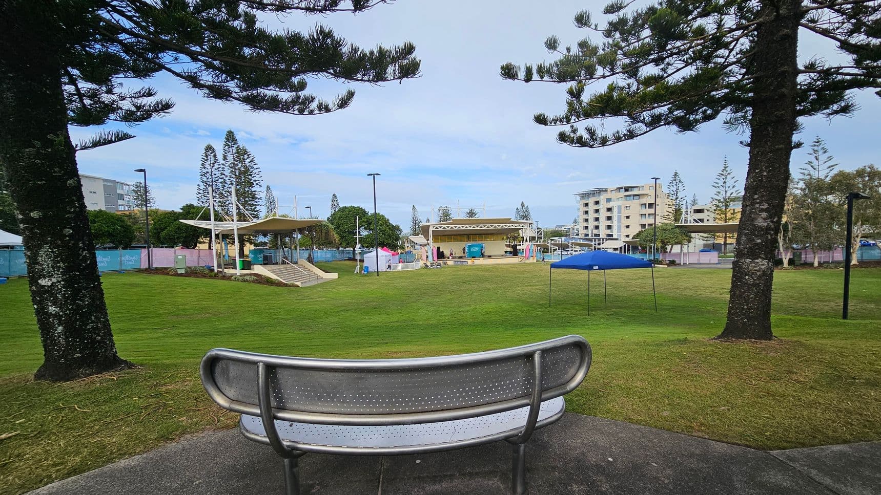 A park scene with a metal bench in the foreground, positioned on a concrete path. Two large trees frame the view on either side. In the middle distance, a grassy area leads to a stage with a canopy and steps. Several tents and structures are scattered around, with a few buildings visible in the background. The area is surrounded by more trees and lampposts.