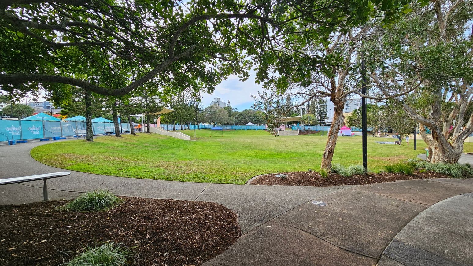 A park with a curved pathway in the foreground, bordered by grass and small plants. On the left, there are blue tents and picnic tables. In the centre, a large grassy area is surrounded by trees. On the right, more trees and a lamppost are visible. The background features additional trees and a glimpse of a building. The scene is framed by overhanging tree branches.
