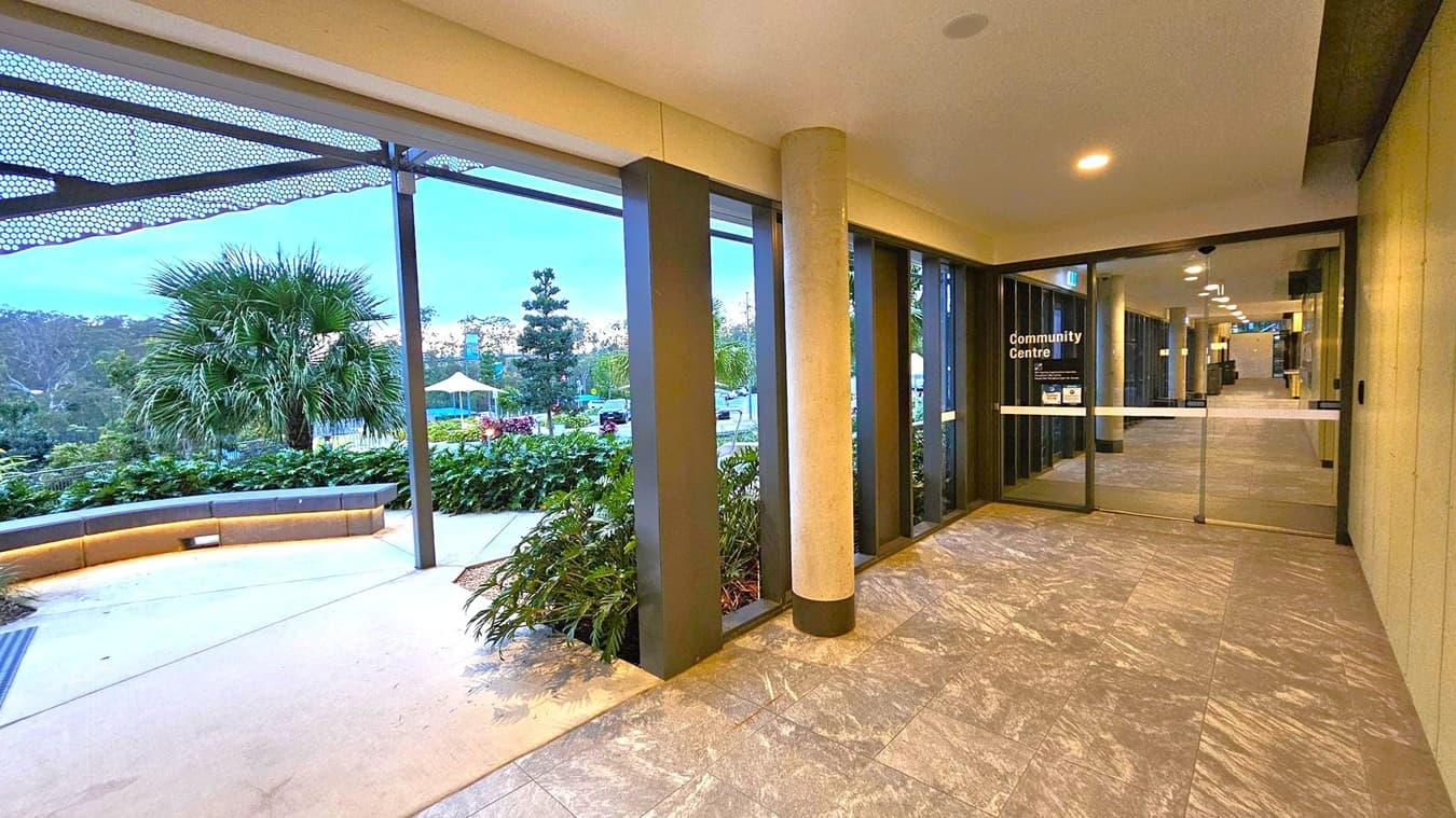 A corridor with a tiled floor leads to a glass door marked "Community Centre" on the right. The ceiling has recessed lighting. On the left, large windows reveal an outdoor area with a palm tree, greenery, and a curved bench. A metal canopy with a perforated design extends above the windows. In the background, there are more trees and a structure with a yellow canopy.