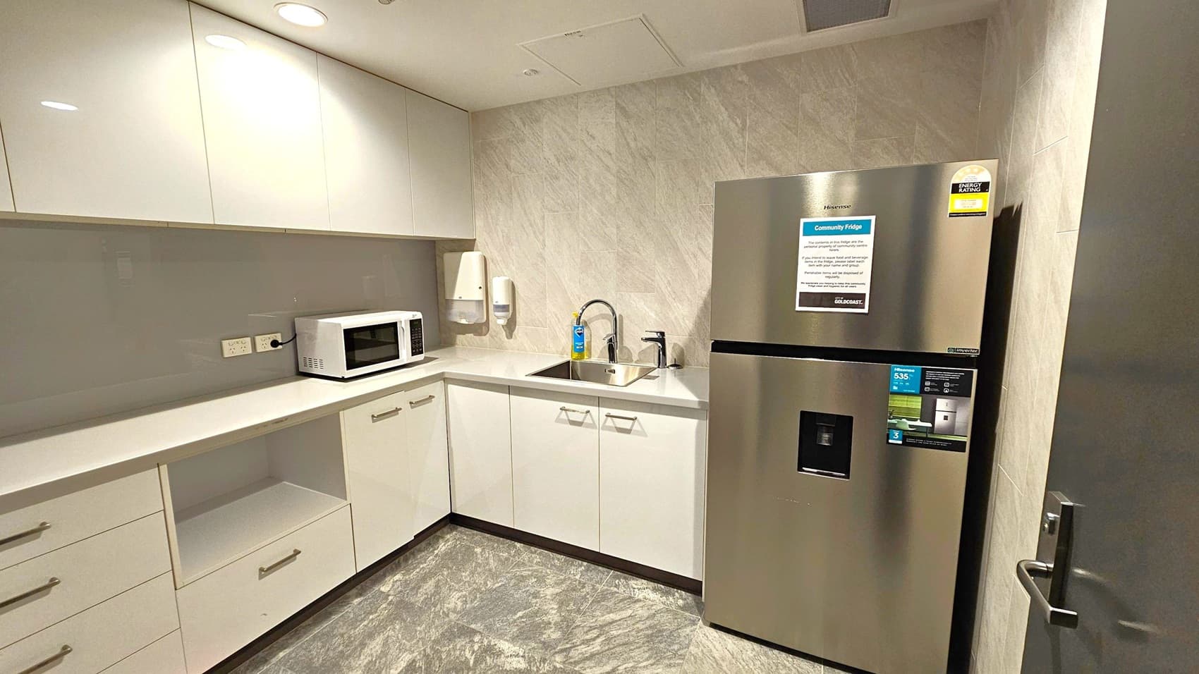A compact kitchen with light-coloured cabinets and a grey marble-patterned floor. On the left, there are upper cabinets and a countertop with a microwave. Below are drawers and an open shelf. In the centre, a sink with a tap is set into the counter, with a soap dispenser nearby. On the right, a stainless steel refrigerator with an energy rating sticker is positioned next to a door. The walls have a light, textured finish, and the ceiling is fitted with recessed lighting.