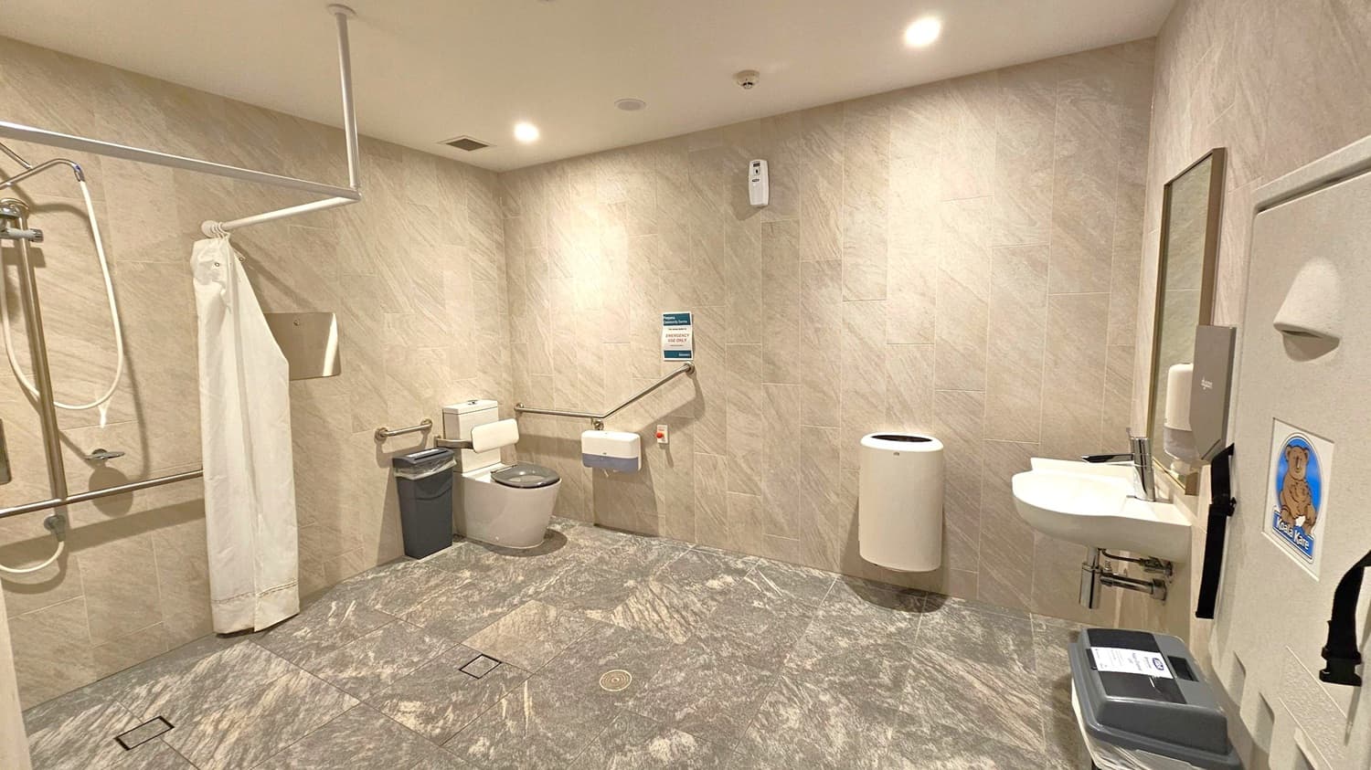 A spacious, accessible bathroom with light grey tiled walls and floor. On the left, a shower area with a white curtain and grab bars. In the centre, a toilet with support rails and a small bin nearby. On the right, a sink with a mirror above it, a soap dispenser, and a baby changing station with a koala logo. The ceiling has recessed lighting, and a white cylindrical bin is positioned against the wall.