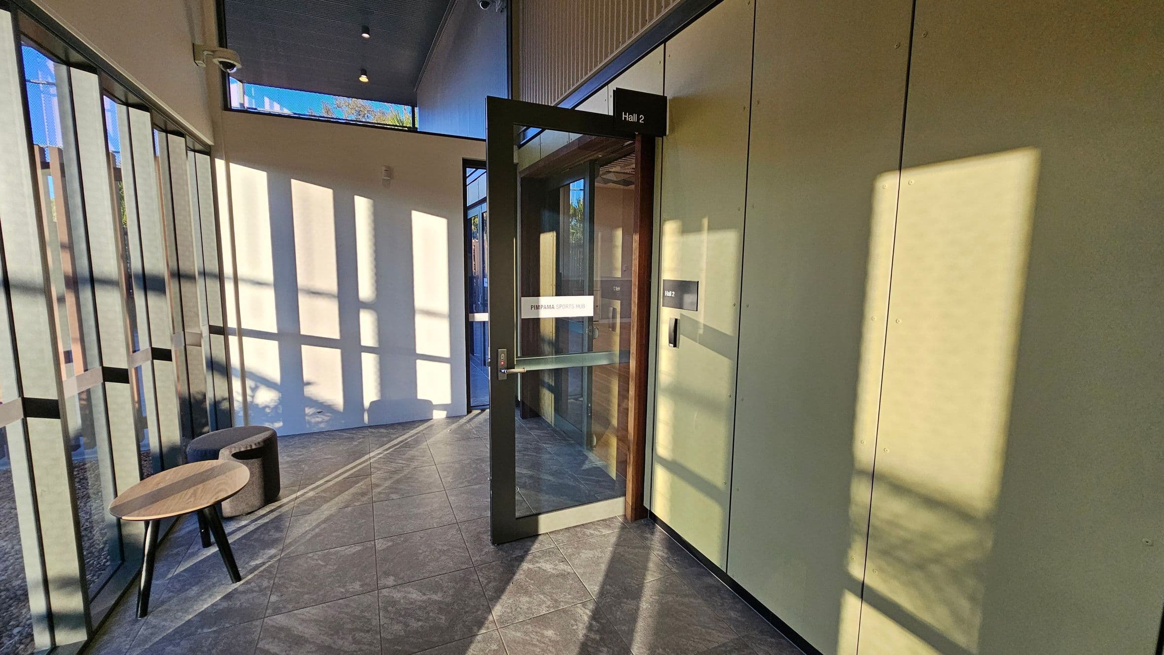 A hallway with a glass door labelled "Hall 2" on the right, leading into a room. The door has a sign reading "Pimpama Sports Hub". The left side features large windows casting shadows on the tiled floor. A small wooden table and a round, cushioned stool are placed near the windows. The ceiling is high with visible light fixtures.