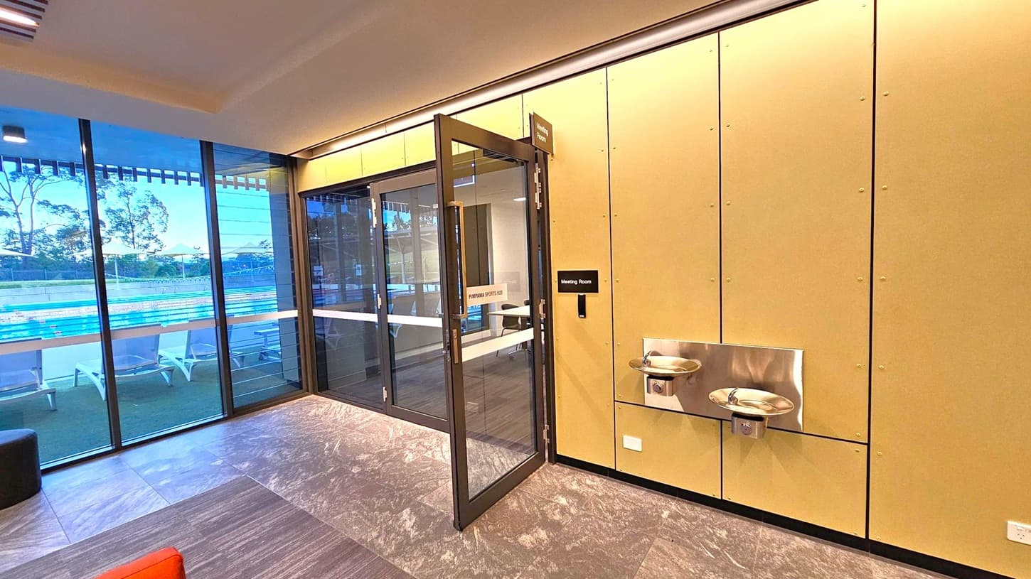 On the left, large glass windows reveal an outdoor swimming pool with lanes and sun loungers. The middle features a glass door leading to a meeting room, with a sign above and on the door. On the right, a beige wall has two stainless steel water fountains. The floor is covered with grey and brown tiles, and the ceiling has recessed lighting.