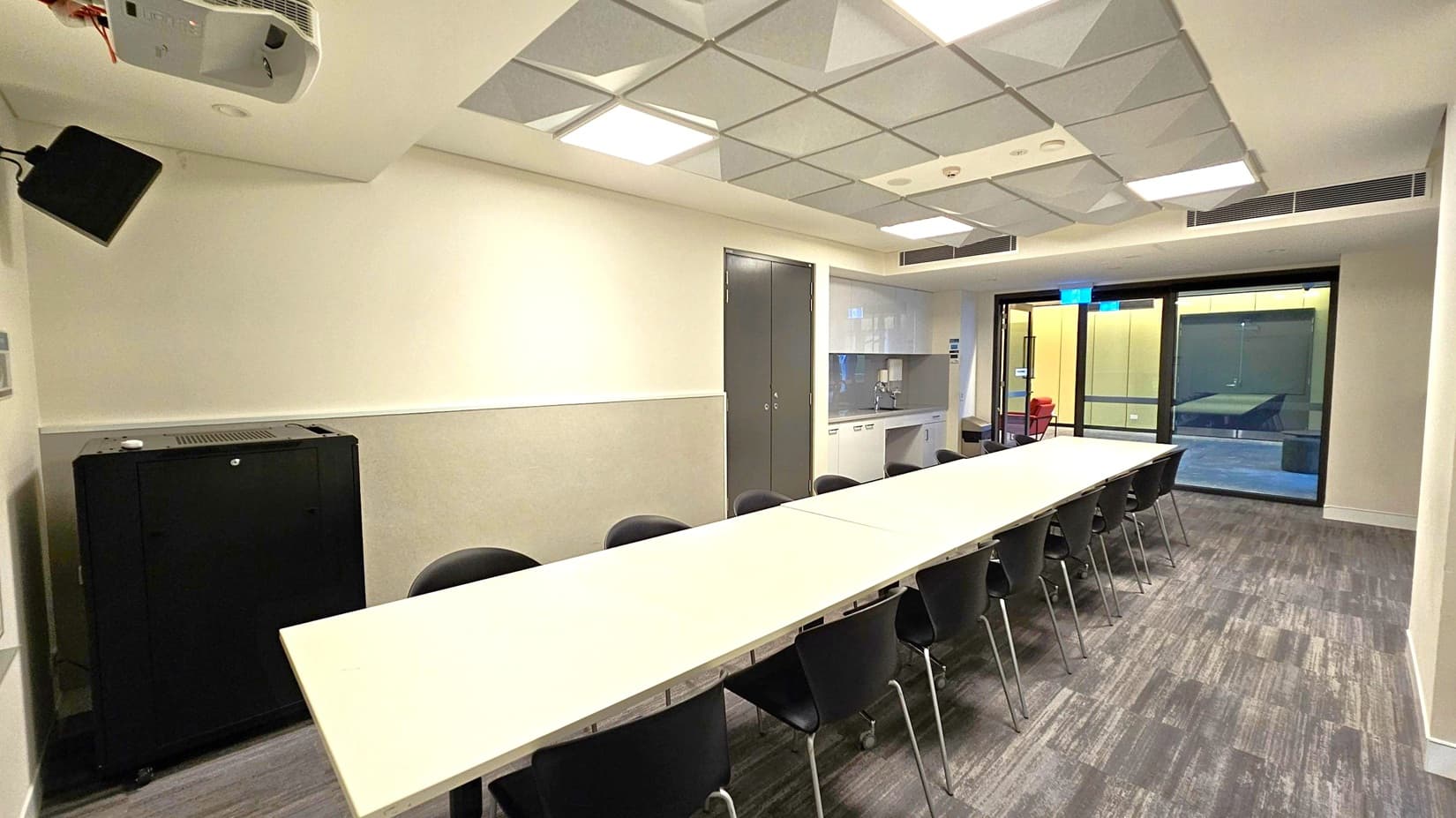 A conference room with a long white table in the centre, surrounded by black chairs. On the left, there's a black cabinet and a wall-mounted speaker. The ceiling has square recessed lights and a grid pattern. On the right, there's a kitchenette with a sink and cabinets, and a glass door leading to another room. The floor is carpeted with a grey and green pattern.
