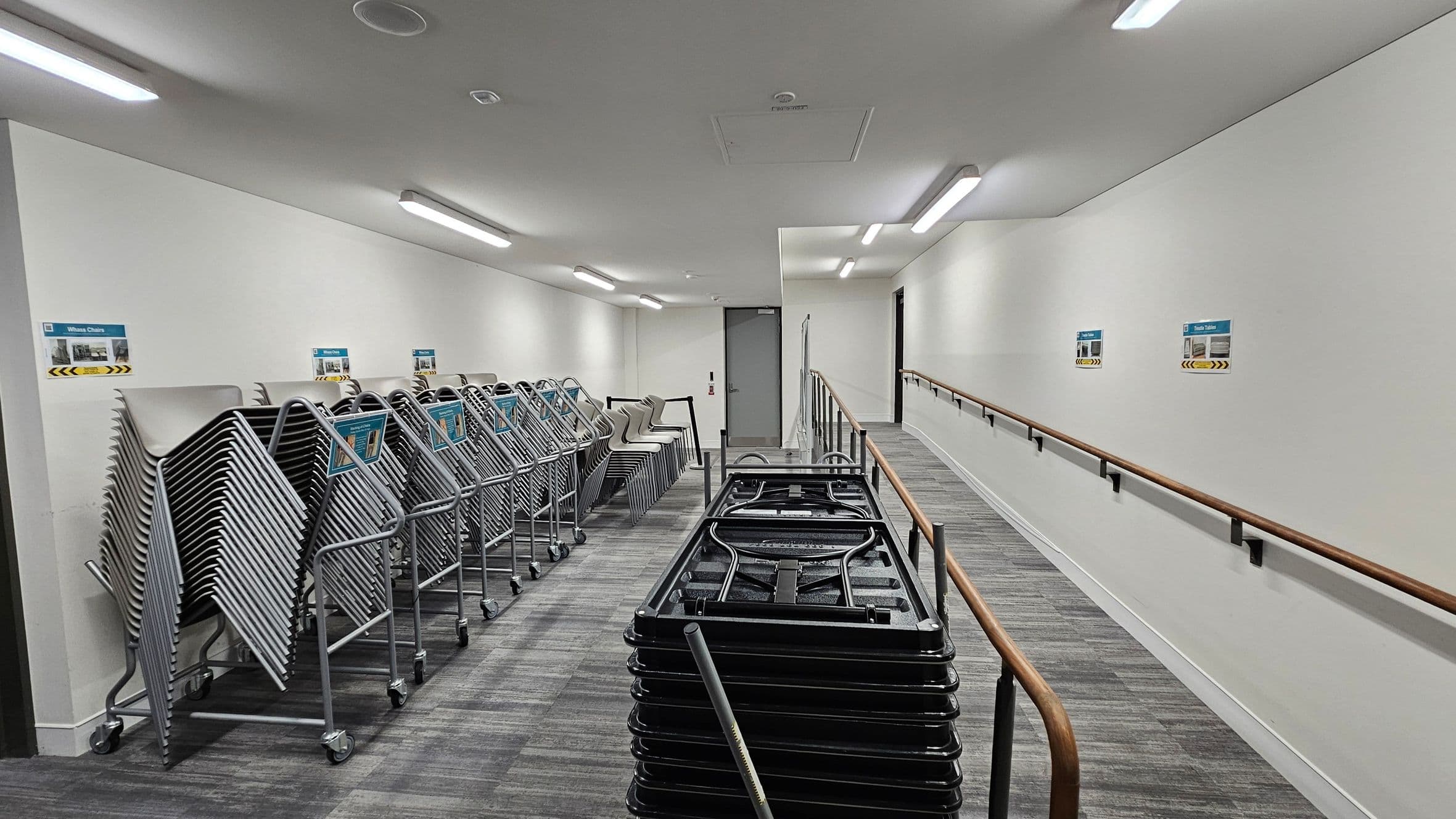 A corridor with a grey floor and white walls. On the left, there are stacks of metal folding chairs on trolleys. On the right, a wooden handrail runs along the wall, with several signs above it. In the centre, a stack of folded tables is visible. The ceiling has several rectangular fluorescent lights. At the end of the corridor, there is a closed door.