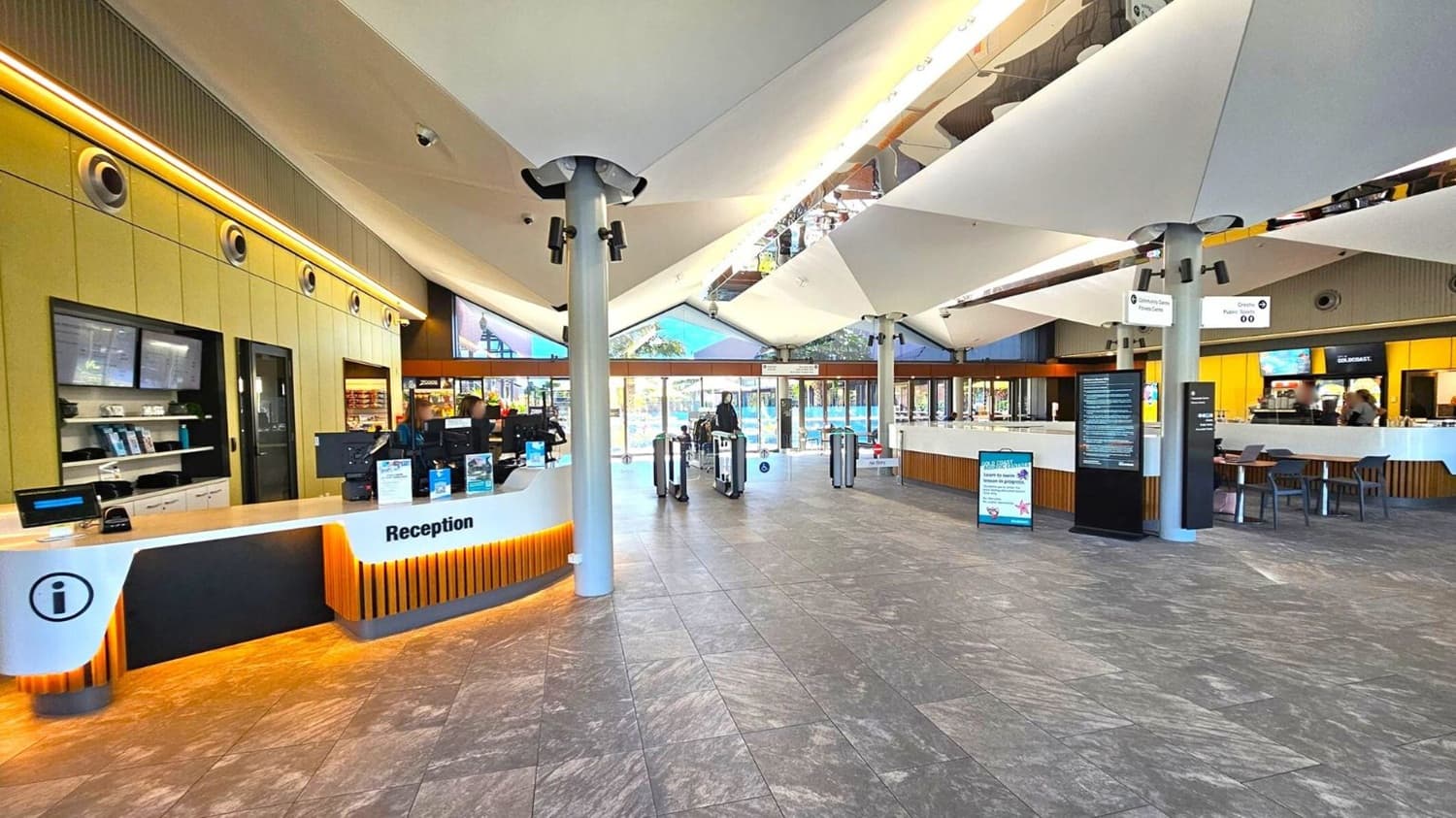 Reception area with a modern design. On the left, a curved reception desk with computers and information signs. Behind it, a wall with built-in shelves and screens. In the centre, turnstiles lead to a glass entrance with views of an outdoor area. On the right, a café area with tables and chairs. The ceiling features large, white, angular panels and the floor is covered with grey tiles.