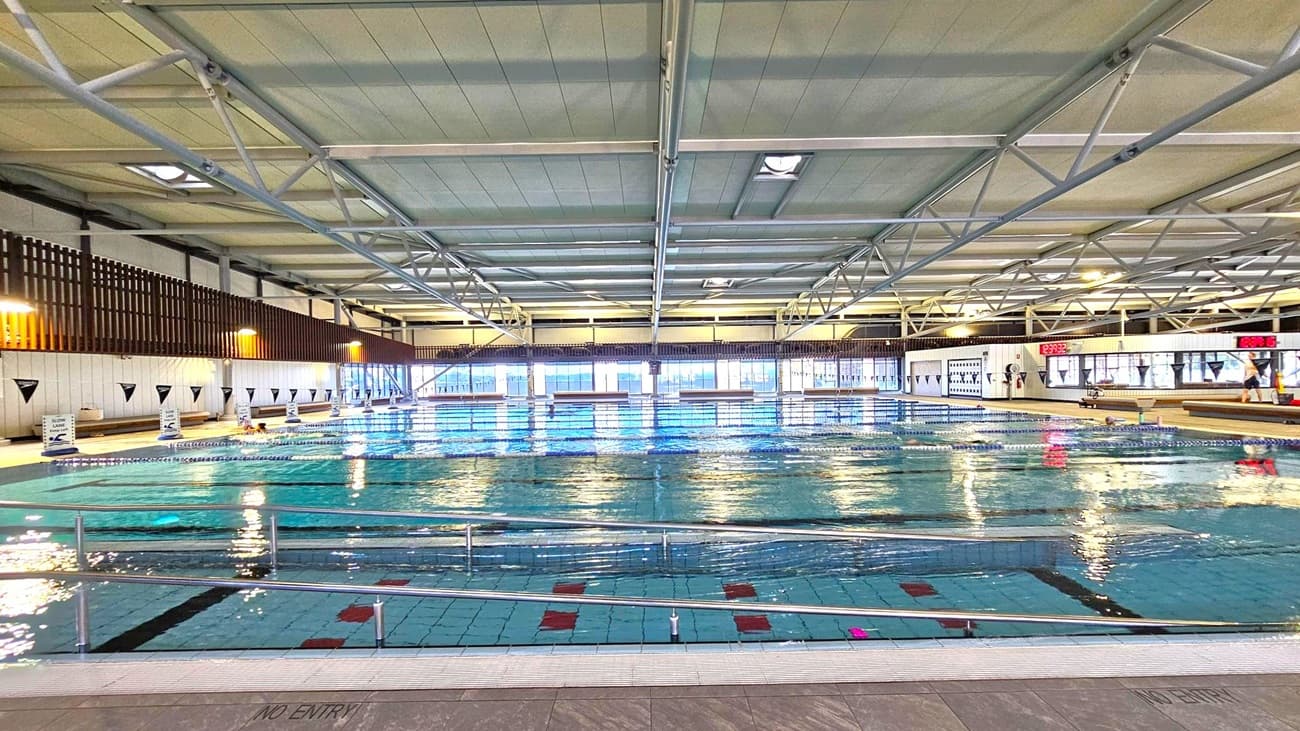 Indoor swimming pool with clear water and lane dividers. The ceiling features metal beams and panels. Large windows line the far wall, allowing natural light to illuminate the space. Digital clocks are mounted on the right wall, displaying the time. The pool deck is tiled, with a "NO ENTRY" sign visible. Safety railings run along the pool's edge. The left wall has vertical wooden slats and signage indicating swimming lanes.