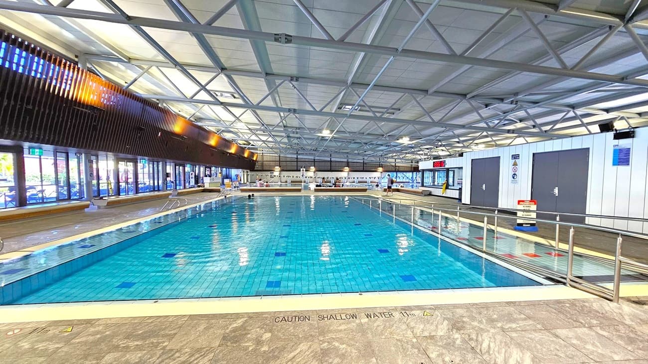 Indoor swimming pool with clear blue water, surrounded by a tiled deck. On the left, large glass doors and windows allow natural light in, with wooden slats above. The right side features white walls with dark doors and safety signs. A digital clock displays the time and temperature. The ceiling has a grid of metal beams and panels. A caution sign on the floor warns of shallow water. A few people are visible near the pool's edge.