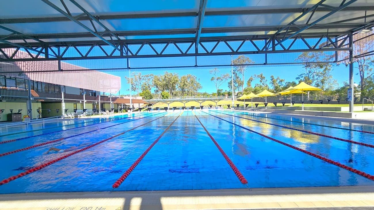 A large outdoor swimming pool with multiple lanes, separated by red lane dividers, stretches into the distance. On the left, a building with a brown roof and glass windows runs parallel to the pool. On the right, several yellow umbrellas provide shade over seating areas. In the background, trees and greenery are visible. The pool is covered by a metal and fabric structure, casting shadows on the water. The floor is tiled at the pool's edge.