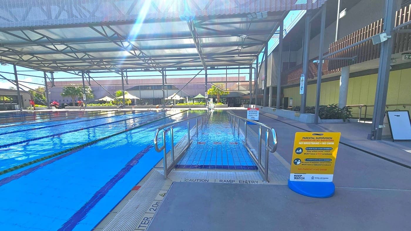 An indoor swimming pool with multiple lanes on the left, separated by lane dividers. In the centre, a ramp with metal railings leads into the water. On the right, a sign with safety instructions is placed on the pool deck. The ceiling is supported by a metal framework, and the background features a building with large windows and some greenery, including palm trees and plants. The floor is tiled near the pool and concrete on the deck.