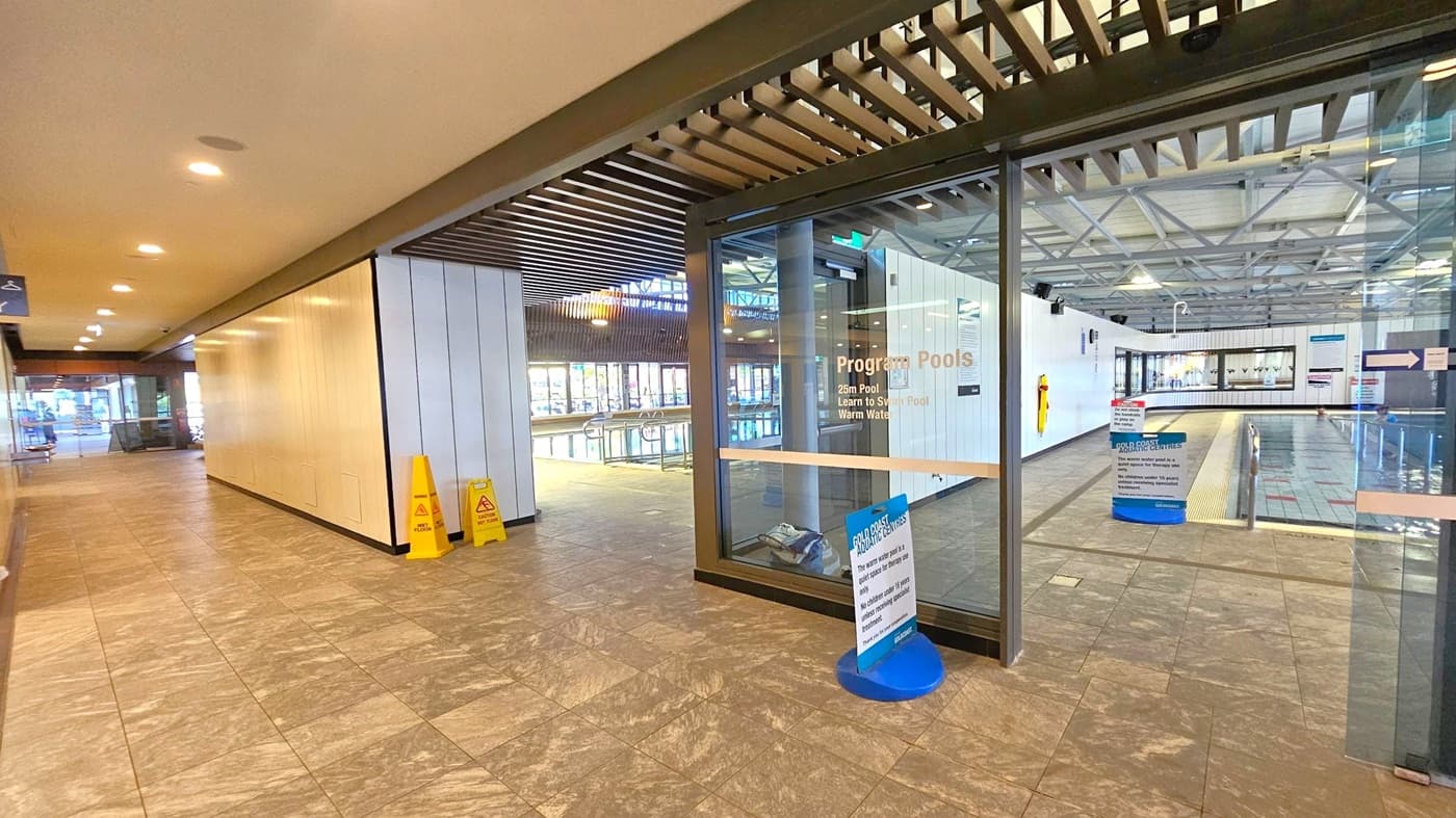 A hallway with a tiled floor and a ceiling with recessed lighting. On the right, there is a glass entrance to a pool area, labelled "Program Pools," with a view of the swimming pool inside. Two yellow caution signs are placed near the entrance. The left side of the hallway has a plain wall and leads to an open area with seating. The pool area has a high ceiling with exposed beams and large windows allowing natural light.