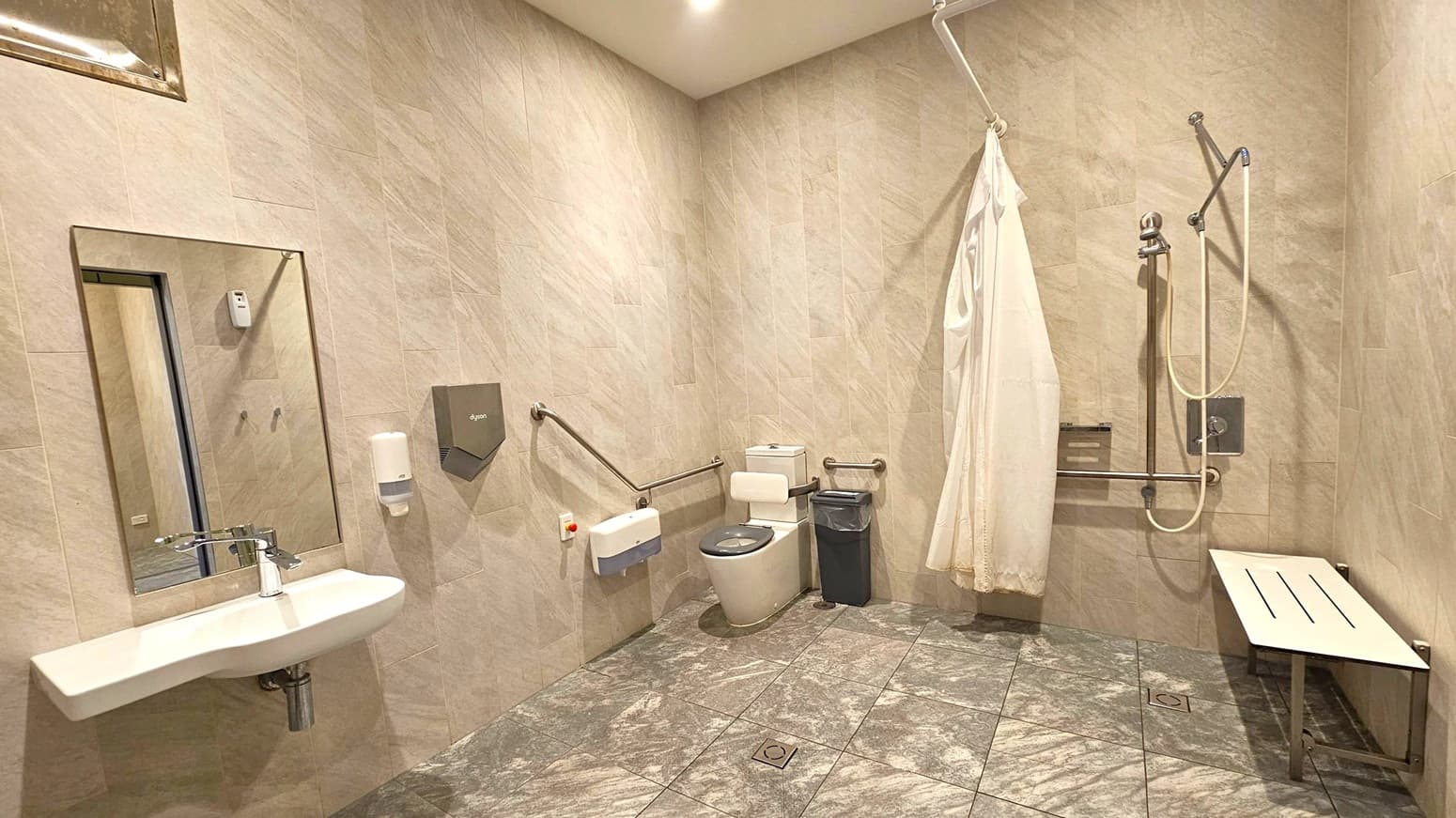 A spacious, accessible bathroom with grey tiled walls and floor. On the left, a wall-mounted sink with a mirror above it, accompanied by a soap dispenser and a hand dryer. In the centre, a toilet with support rails on both sides and a small bin nearby. On the right, a shower area with a curtain, handheld showerhead, and a foldable bench. The ceiling is plain with recessed lighting.