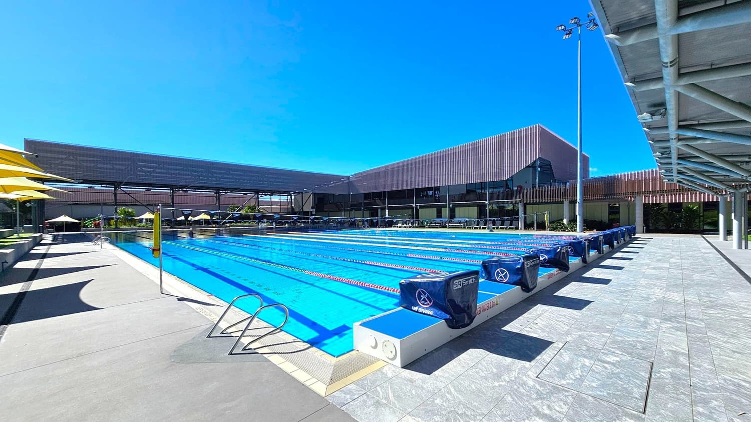 An outdoor swimming pool with multiple lanes, each marked by red and white lane dividers. On the left, there are yellow umbrellas providing shade over seating areas. The right side features a tiled walkway with a metal railing and a covered structure overhead. In the background, modern buildings with a mix of glass and metal elements are visible, along with more seating and umbrellas. The pool area is well-lit with tall floodlights.