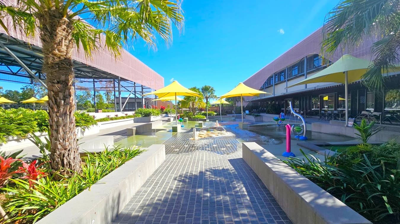 A courtyard with a paved walkway in the centre, flanked by low concrete walls and lush green plants. Yellow umbrellas provide shade over seating areas on both sides. On the left, a structure with a metal framework and a pink mesh roof is visible. On the right, a building with large windows and a shaded patio area. In the middle, water features and play equipment are present. Palm trees and other tropical plants are scattered throughout the area.