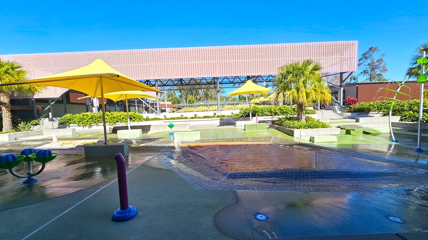 A water play area with a wet surface and various water features, including a purple pipe and blue and green structures. Yellow umbrellas provide shade, and there are palm trees and lush greenery around. In the background, a large structure with a slatted roof and metal framework is visible, along with steps leading up to it. The area is surrounded by landscaped plants and shrubs.