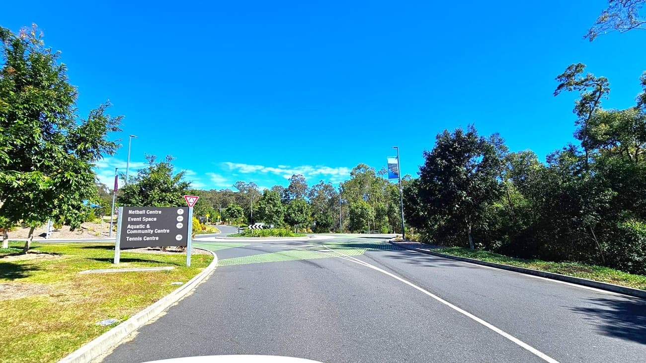 A road with a roundabout in the centre, surrounded by trees and greenery. On the left, a sign lists directions to various centres, including a netball centre and aquatic centre. The road has a green patterned section near the roundabout. On the right, a tall streetlight and a banner are visible. The area is bordered by dense trees and shrubs.