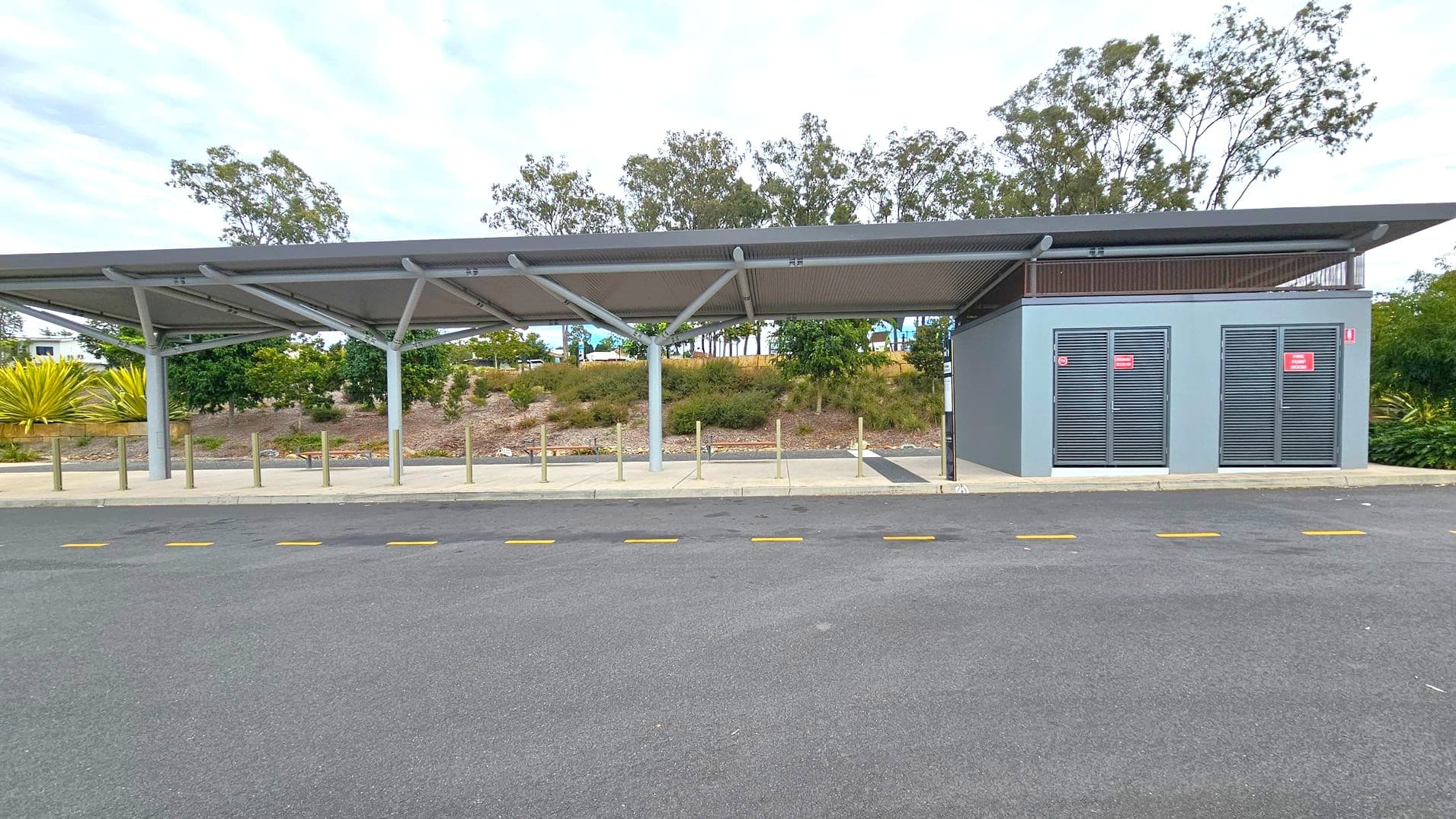 A bus stop with a modern shelter featuring a slanted roof supported by metal beams. On the right, there is a grey utility building with two vented doors displaying red signs. The foreground has a paved area with yellow dashed lines on the road. In the background, there are trees and shrubs on a slightly elevated grassy area. The sky is overcast.