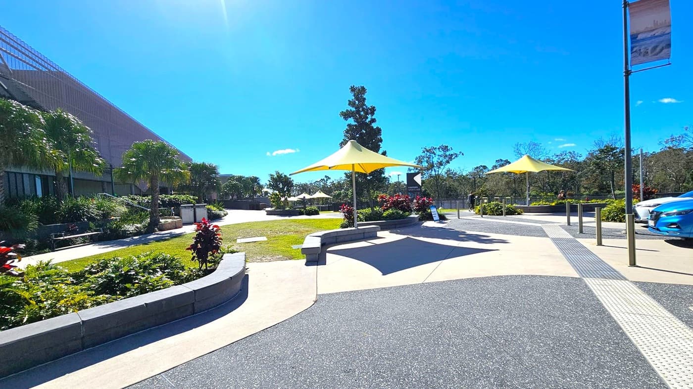 A paved outdoor area with yellow triangular shade structures in the centre and right. The left side features a large building with a slanted roof, surrounded by lush greenery and palm trees. The foreground has a curved stone bench with plants. In the background, there are more trees and a clear blue sky. A blue car is partially visible on the right, and a tall pole with a banner is near the car.