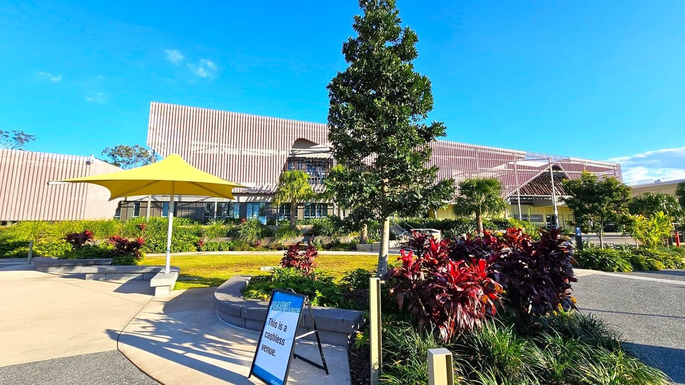 A modern building with a textured facade is in the background, surrounded by lush greenery and various plants. In the foreground, a yellow umbrella provides shade over a seating area. A sign on the left reads "This is a cashless venue." The ground is a mix of concrete and gravel paths, and a small tree is centrally located, adding to the landscaped environment.