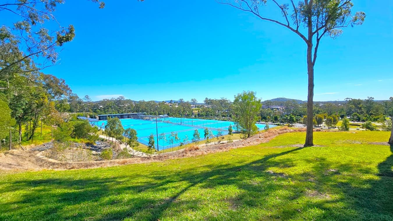 A grassy hill in the foreground with a tall tree on the right. In the middle, several bright blue tennis courts are visible, surrounded by a fence. To the left, there are more trees and a path. The background features more trees and a distant view of hills and buildings.