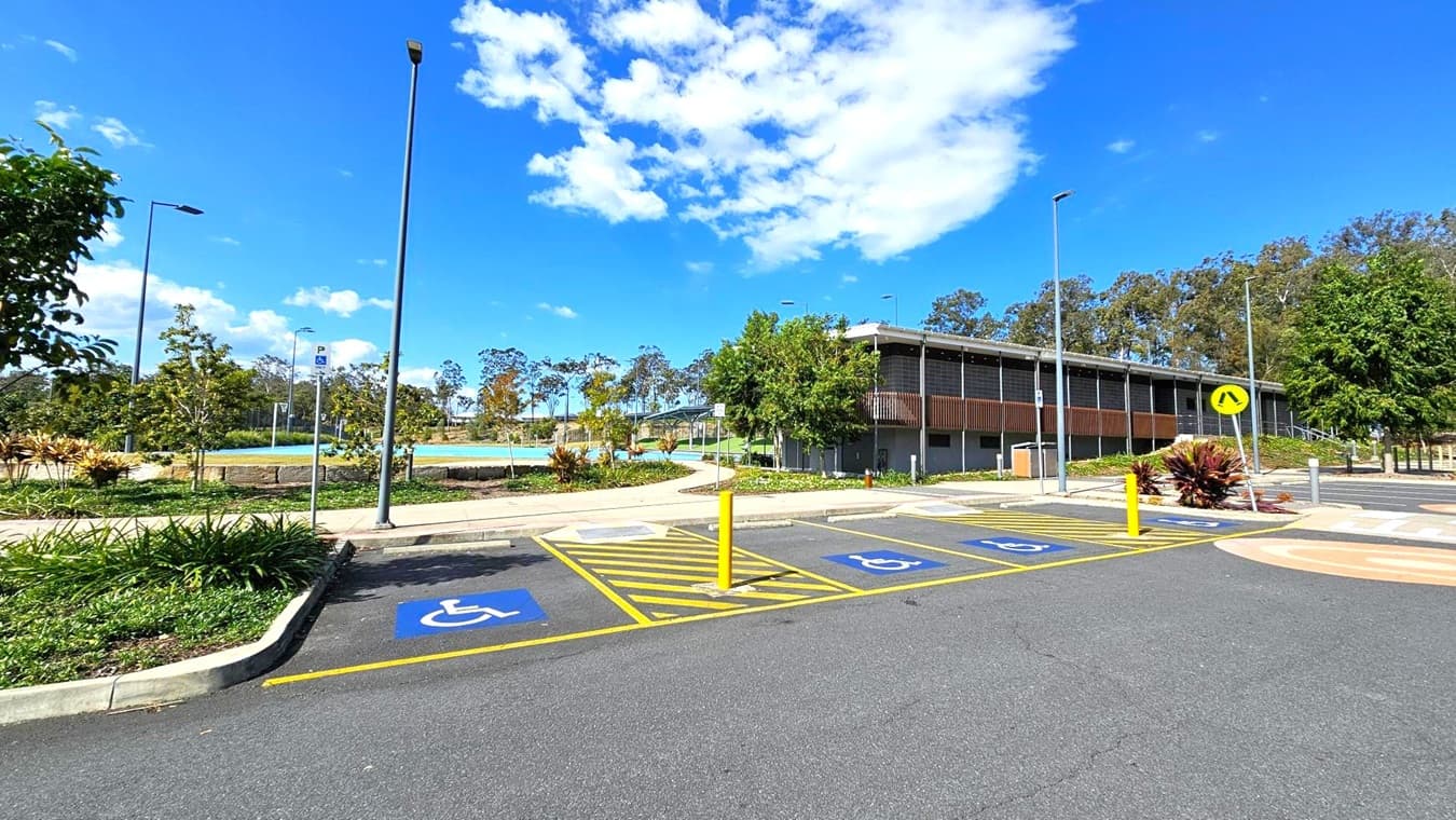 A parking area with designated disabled parking spaces marked in blue on the asphalt. Yellow lines and a bollard separate the spaces. On the right, a modern building with a flat roof and wooden slats is partially obscured by trees. In the background, a landscaped area with small trees and a pathway is visible. Several tall streetlights are scattered throughout the scene.