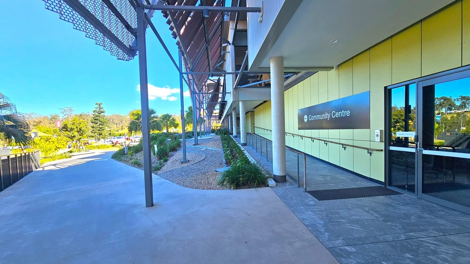 A pathway leads to a modern building on the right, marked as a "Community Centre" with a sign. The building features a glass entrance and a series of vertical columns. On the left, there is a landscaped area with small plants and trees, bordered by a metal fence. Overhead, a metal awning with a geometric pattern provides shade. The ground is paved with concrete, and the area is well-lit by natural light.