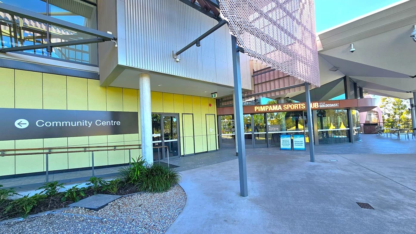 Modern building exterior with a sign pointing left to the "Community Centre" on the left side. The wall is clad in yellow panels with a glass door entrance. In the centre, a cylindrical column supports the structure. On the right, the entrance to "Pimpama Sports Hub" is visible, with large glass windows and a sign above. The foreground features a concrete path and landscaped area with small plants and gravel. The ceiling has a geometric design with metal and glass elements.