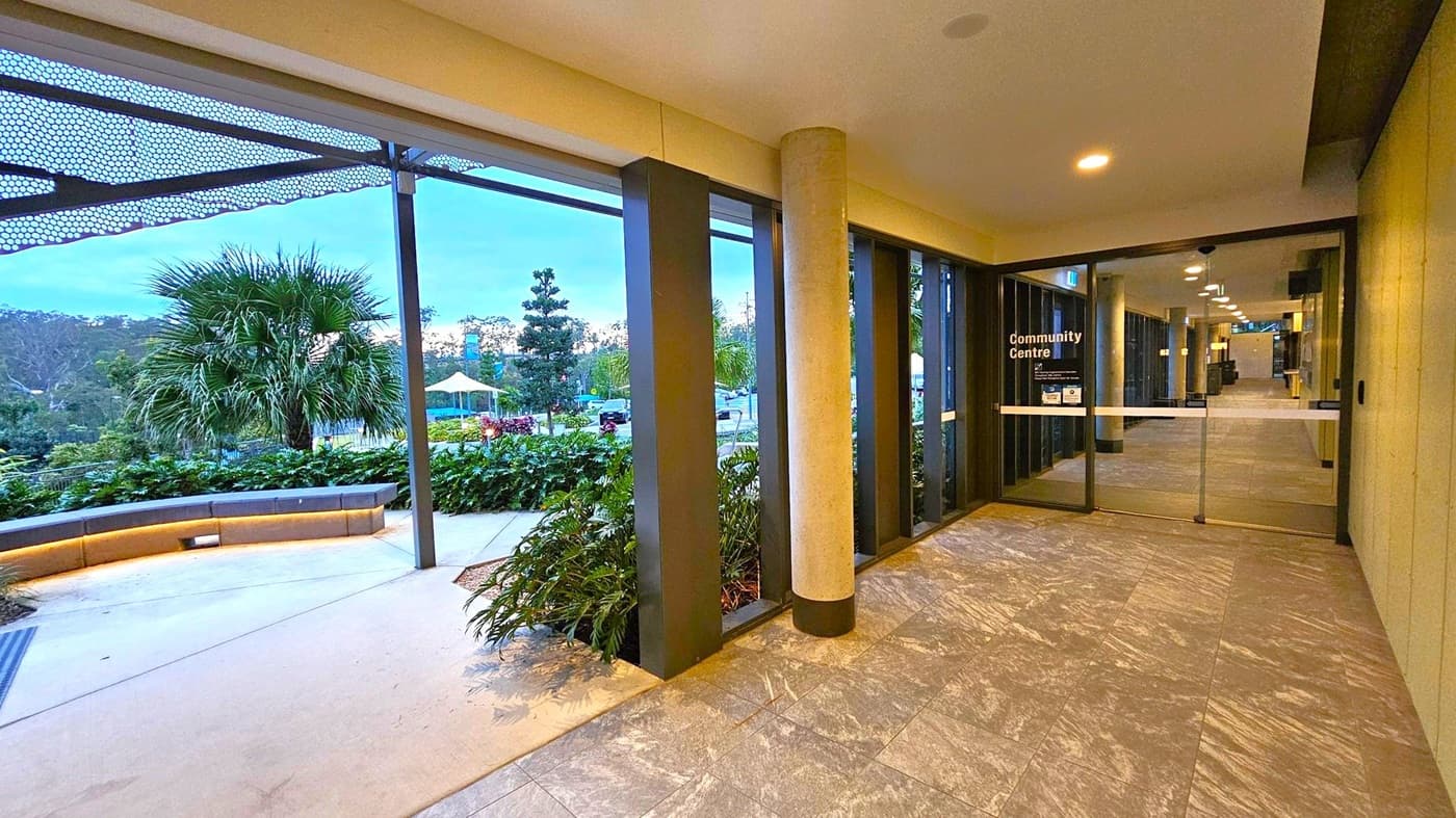 A corridor with a tiled floor leads to a glass door marked "Community Centre" on the right. The ceiling has recessed lighting. On the left, large windows reveal an outdoor area with a curved bench, lush greenery, and a palm tree. A metal canopy with a perforated design extends overhead. In the background, there are more trees and a structure with a yellow canopy.