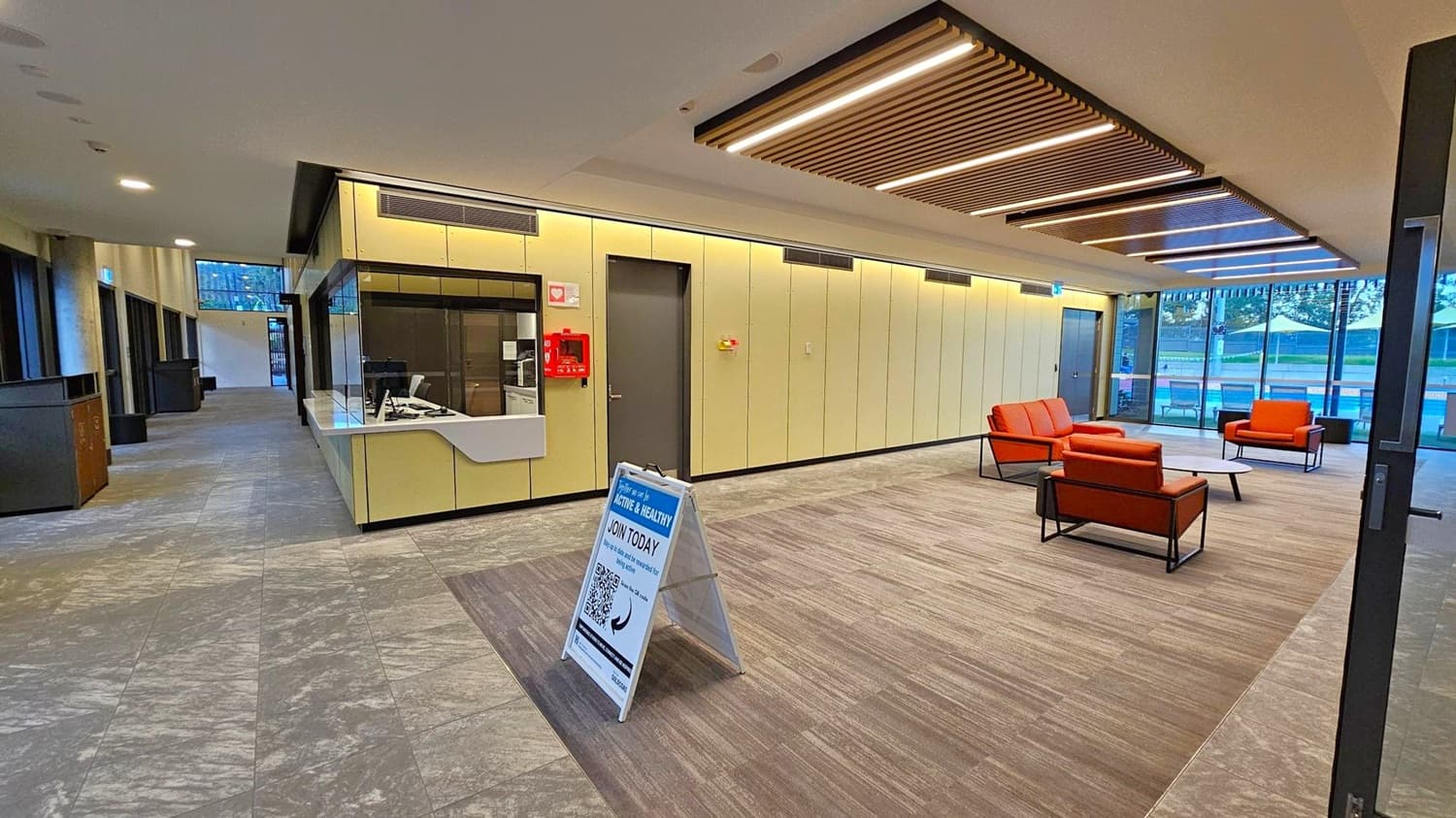 A modern interior space with a reception area on the left featuring a counter and a small window. A signboard stands in front of it. The right side has a seating area with orange chairs and a low table on a carpeted section. Large windows in the background reveal an outdoor area with tables and umbrellas. The ceiling has wooden slats with integrated lighting, and the floor is a mix of tiled and carpeted surfaces.