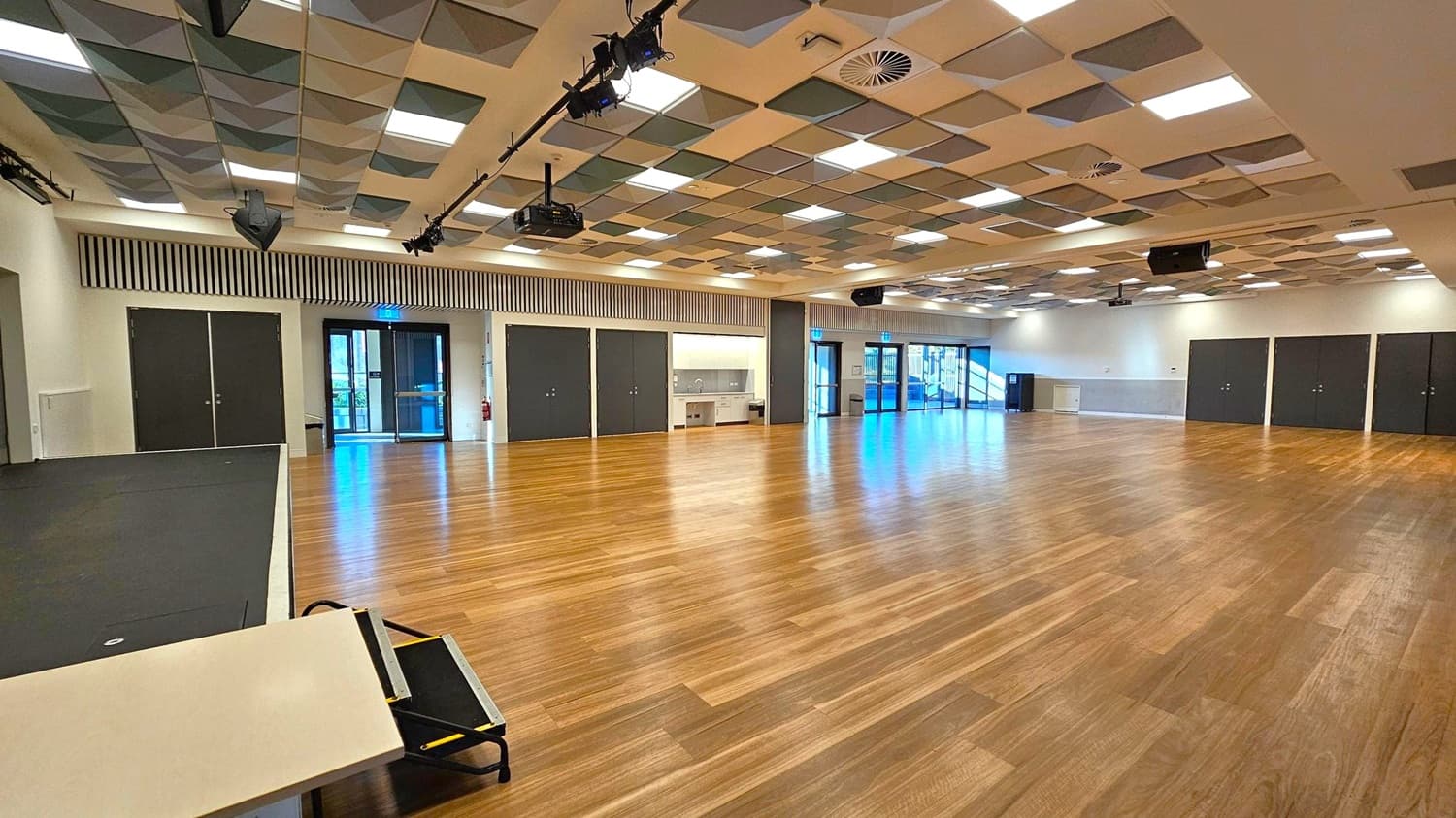 A spacious room with a wooden floor and a patterned ceiling featuring square panels in various shades. On the left, there is a small stage area with a black surface and a table with a folded chair. The back wall has several black double doors and a kitchenette area with a sink. Large glass doors on the right lead outside, allowing natural light to enter. The ceiling has lighting fixtures and speakers, with a projector mounted near the centre.