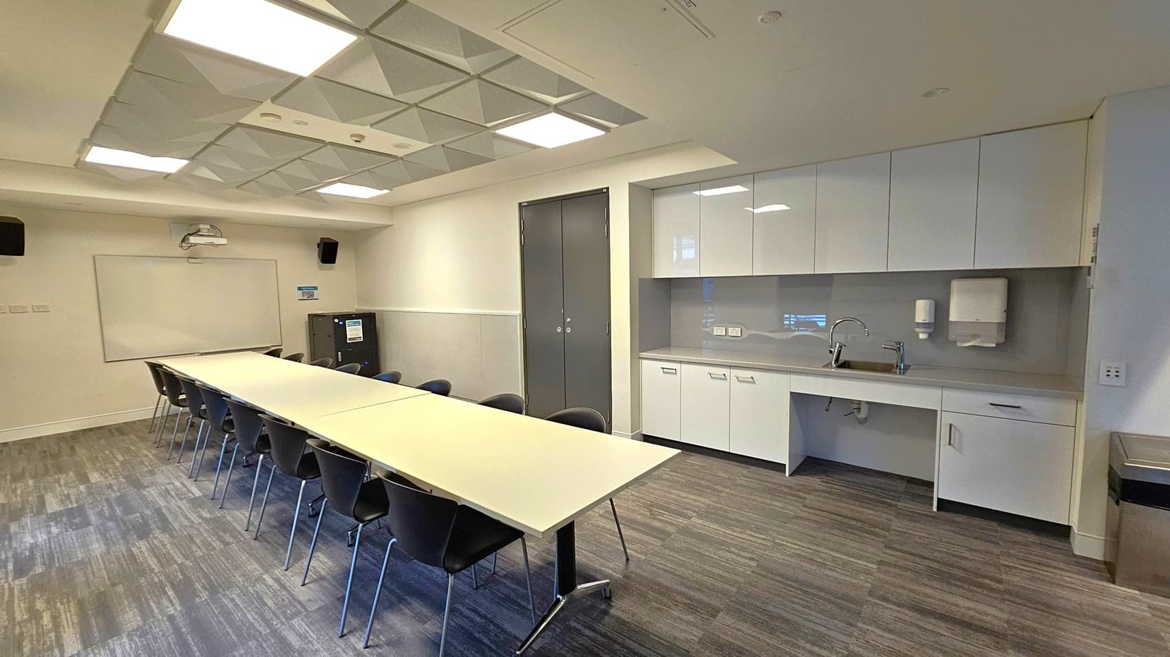 A modern conference room with a long rectangular table in the centre, surrounded by black chairs. On the right, there is a kitchenette with white cabinets, a sink, and a paper towel dispenser. The back wall features a whiteboard and a projector. The ceiling has recessed lighting and a grid pattern. The floor is carpeted with a mix of grey and green tones.