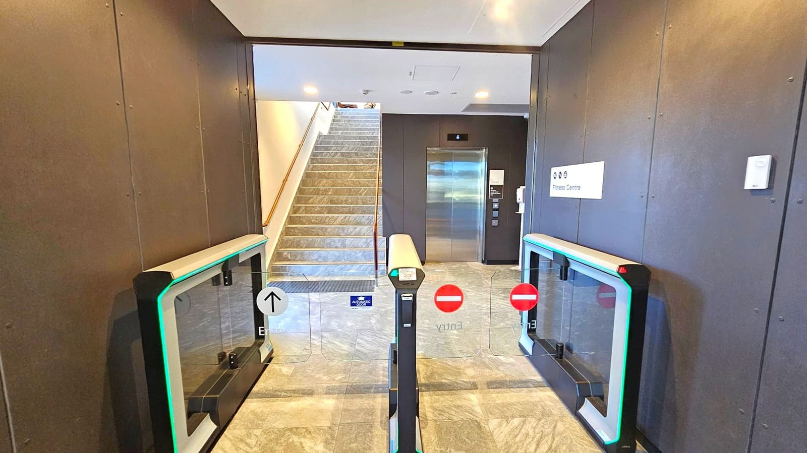A modern entrance area with a turnstile gate in the foreground, featuring glass panels and green LED lights. The floor is tiled with a grey pattern. In the background, a staircase with a metal handrail leads upwards on the left, while a lift with a metallic door is on the right. A sign on the wall indicates a "Fitness Centre." The ceiling is white with recessed lighting.