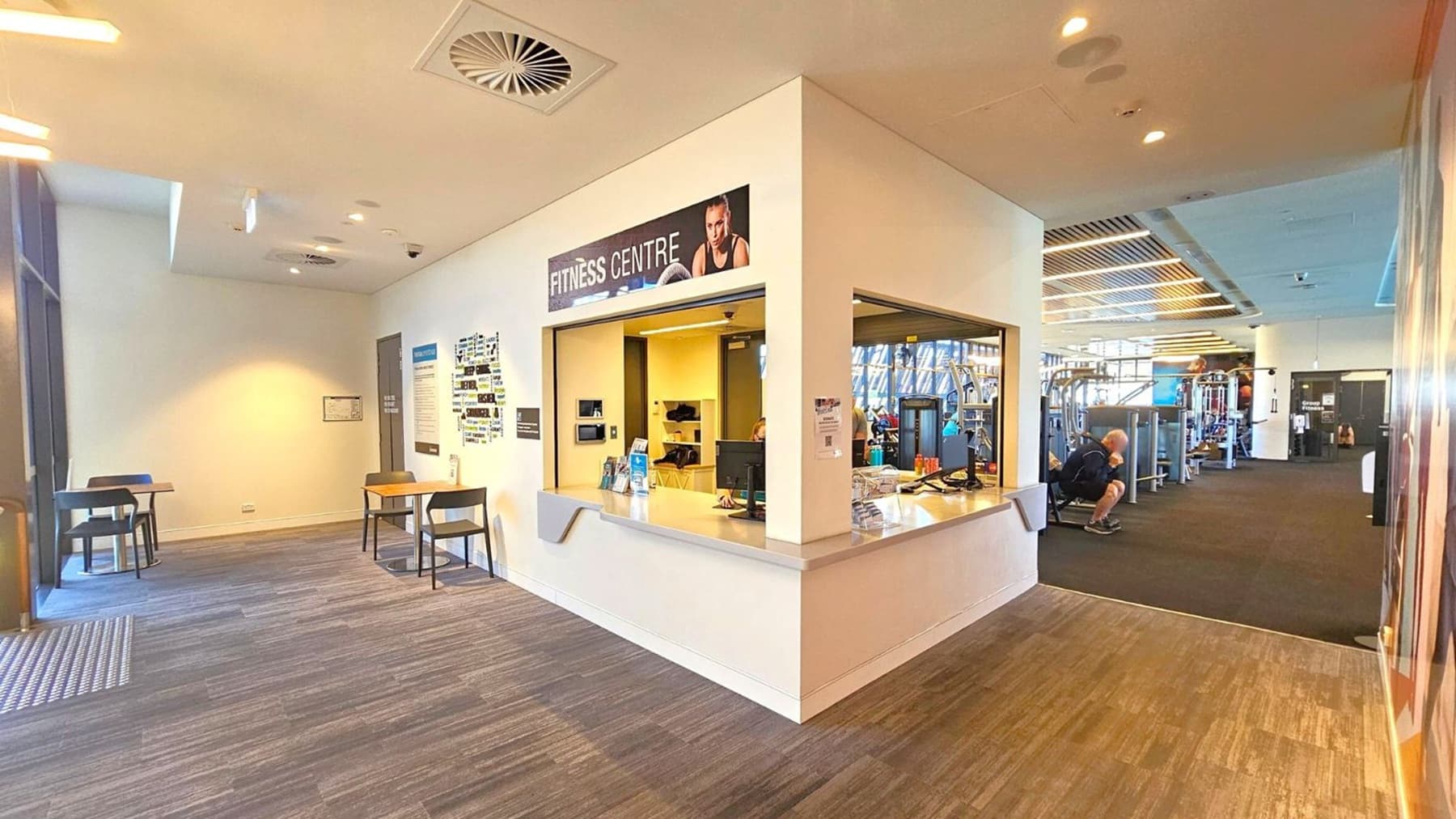 A fitness centre with a reception area in the middle, featuring a counter with a computer and promotional materials. To the left, there are small tables and chairs against a light-coloured wall. The floor is a mix of grey and purple tones. On the right, gym equipment and a person exercising are visible. The ceiling has recessed lighting and ventilation panels.
