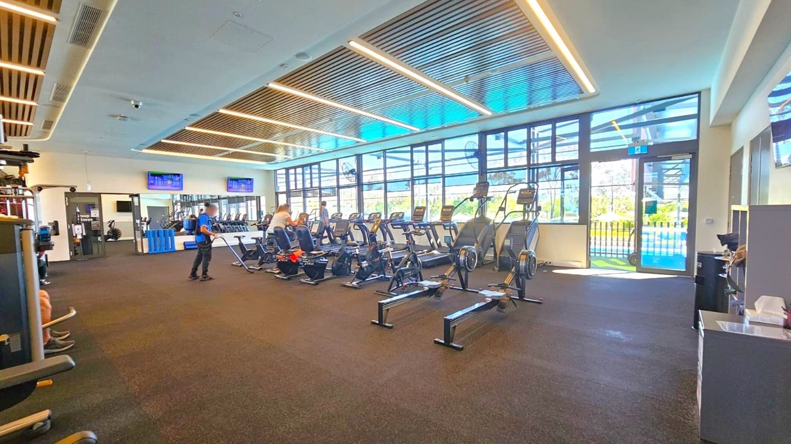 A spacious gym interior with a dark carpeted floor and a ceiling featuring wooden slats and recessed lighting. On the right, there are several rowing machines and exercise bikes lined up facing large windows that let in natural light. In the middle, a person is using a rowing machine, while another stands nearby. On the left, there are various gym equipment and a doorway. Two wall-mounted screens are visible above the equipment.