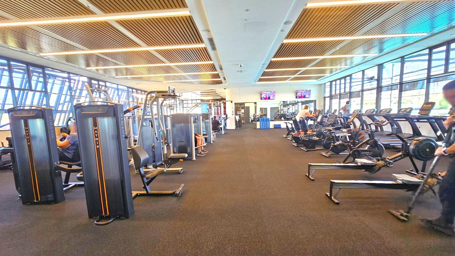 A spacious gym with a variety of exercise equipment. On the left, there are several weight machines with people using them. In the middle, the floor is covered with dark carpeting, and there are two televisions mounted on the wall. On the right, there are treadmills and rowing machines, with a few people exercising. The ceiling features wooden slats with integrated lighting, and large windows line the right side, allowing natural light to enter.