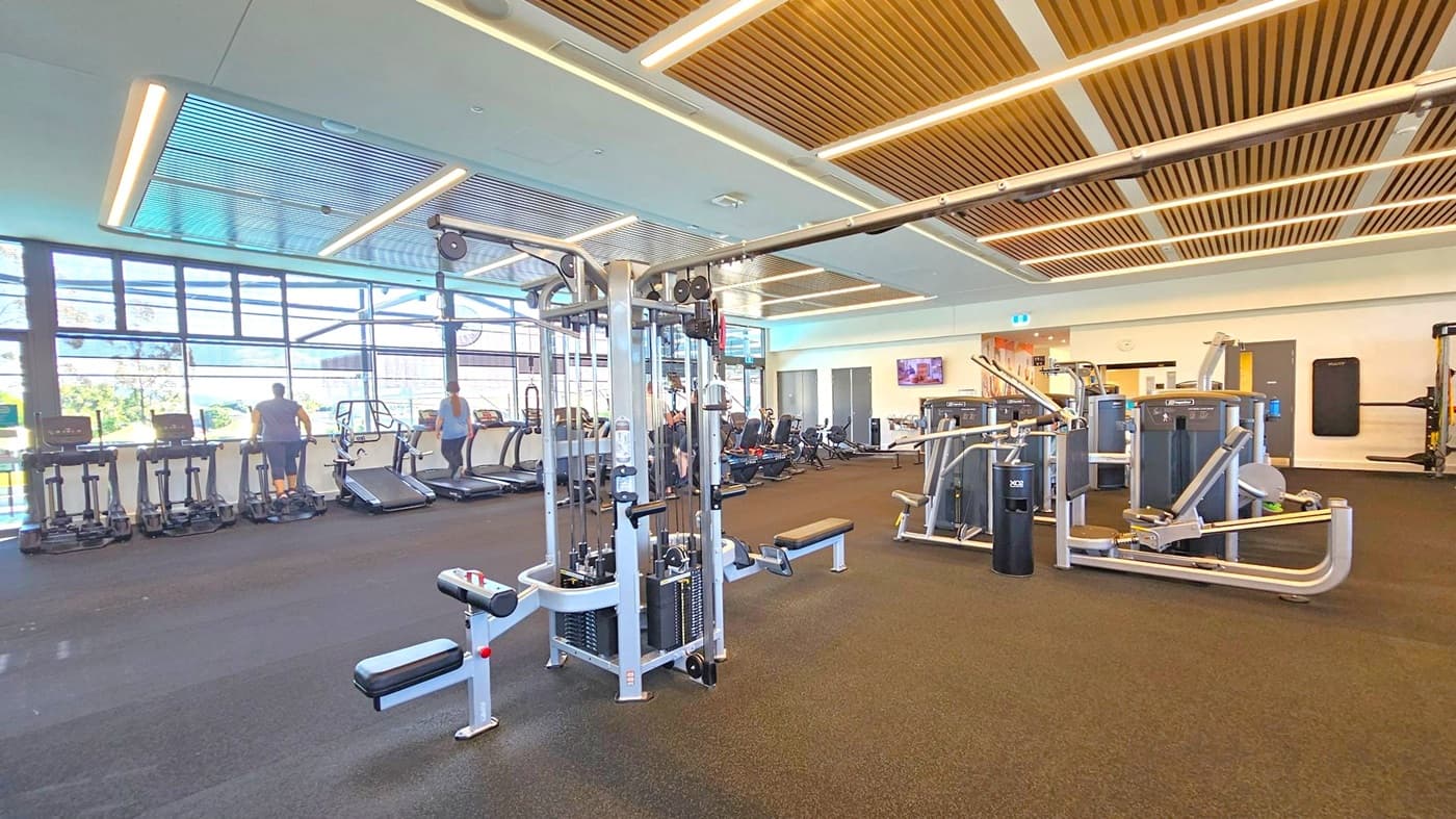 A spacious gym with a variety of exercise equipment. On the left, several treadmills are positioned near large windows, with two people using them. In the centre, a multi-functional weight machine is visible. To the right, there are more weight machines and exercise equipment. The floor is covered with dark rubber matting, and the ceiling features wooden slats with integrated lighting. The background includes a wall-mounted TV and doors.