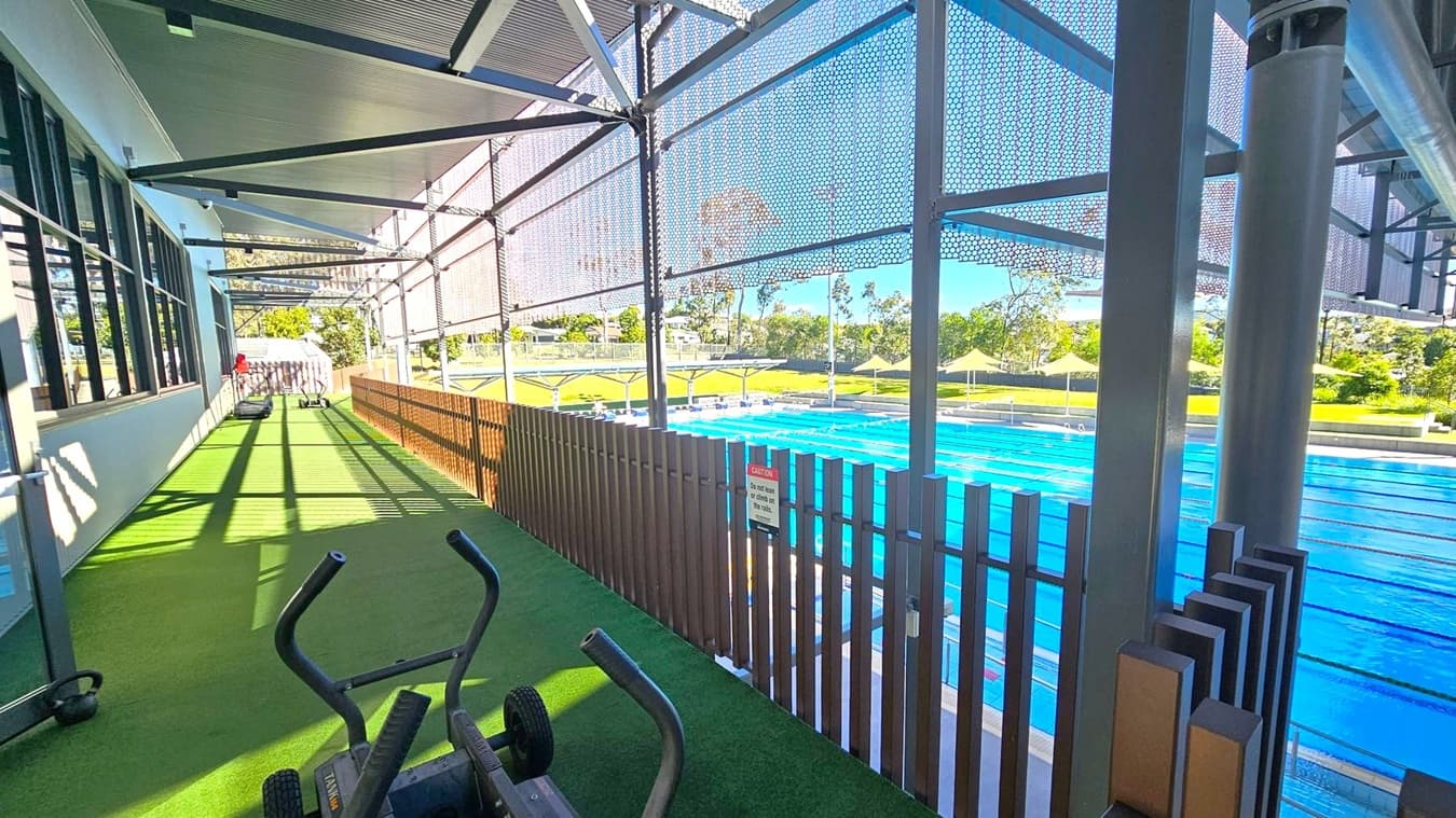 A gym area with artificial grass flooring is on the left, featuring exercise equipment like a treadmill and a stationary bike. Large windows line the left wall. On the right, a metal fence separates the gym from an outdoor swimming pool with clear blue water. Yellow sunshades and green grass are visible beyond the pool. The ceiling is made of metal beams and panels, and the area is partially covered by a mesh screen. Trees and a clear sky are in the background.