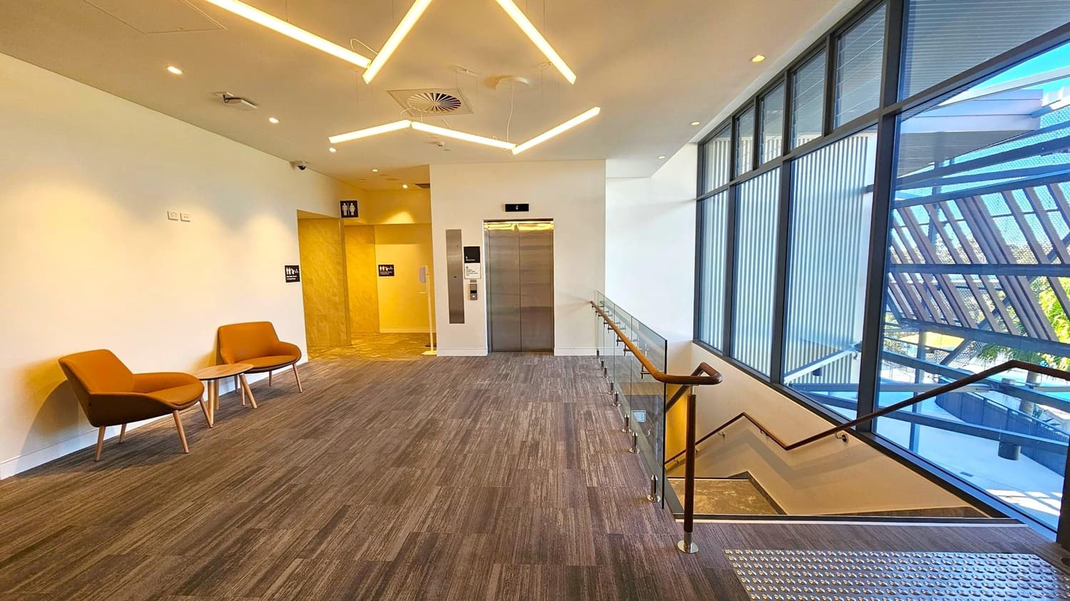 A modern interior space with a grey carpeted floor. On the left, two brown chairs with a small round table are set against a white wall. In the centre, a lift with metallic doors is flanked by signs for restrooms. On the right, a staircase with a wooden handrail descends alongside large windows with a view of an exterior structure. The ceiling features geometric light fixtures and recessed lighting.