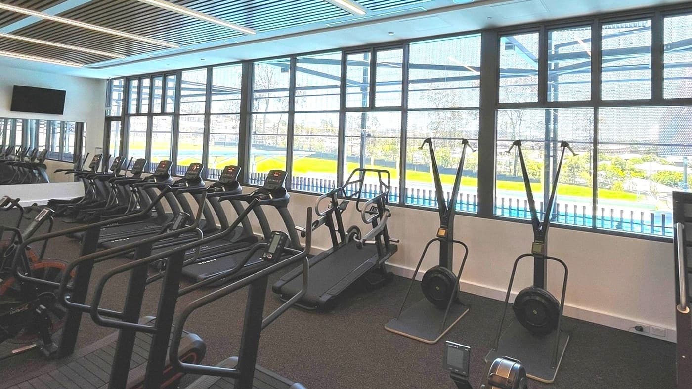A modern gym interior with a row of treadmills on the left and ski machines in the centre. Large windows on the right provide a view of a green outdoor area and a swimming pool. The ceiling features a grid pattern with integrated lighting. The floor is covered with dark carpeting. A wall-mounted TV is visible on the left.
