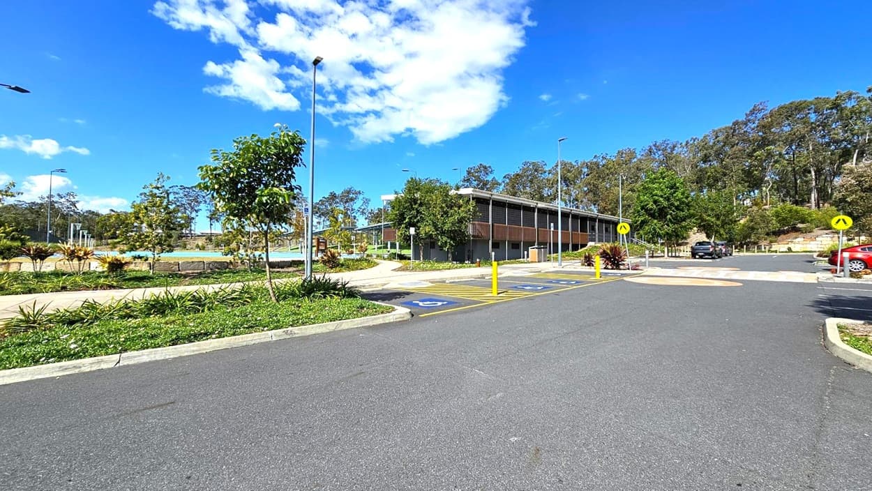 A paved parking area with marked spaces, including disabled parking, is in the foreground. On the left, there are landscaped gardens with small trees and shrubs. In the centre, a modern building with a flat roof and wooden accents is visible. To the right, more parking spaces and a pedestrian crossing are present. The background features a line of tall trees under a clear blue sky with some clouds.