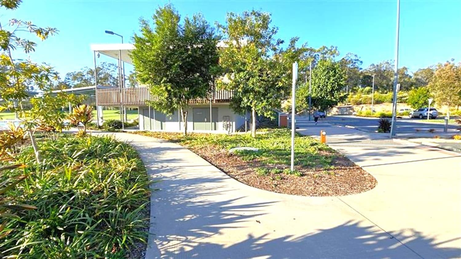 A paved pathway curves through a landscaped area with green shrubs and small trees on both sides. In the background, a modern building with a flat roof and vertical slats is partially visible. The area is well-maintained, with a streetlamp and a few cars parked in the distance. The sky is clear, and the shadows suggest it is a sunny day.